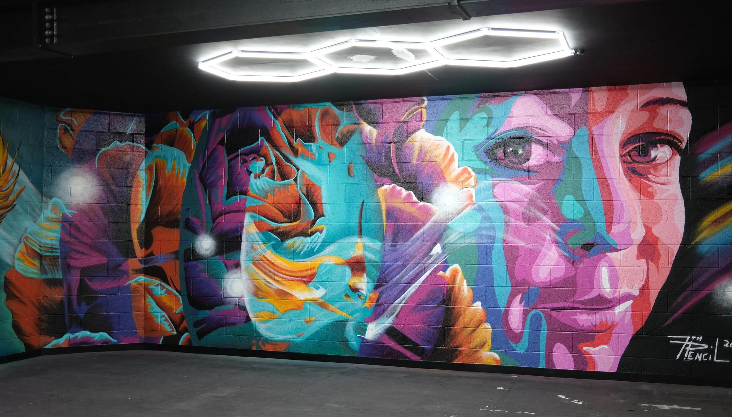 Colorful mural of a woman's face and a lion on a brick wall, with neon ceiling lights above.