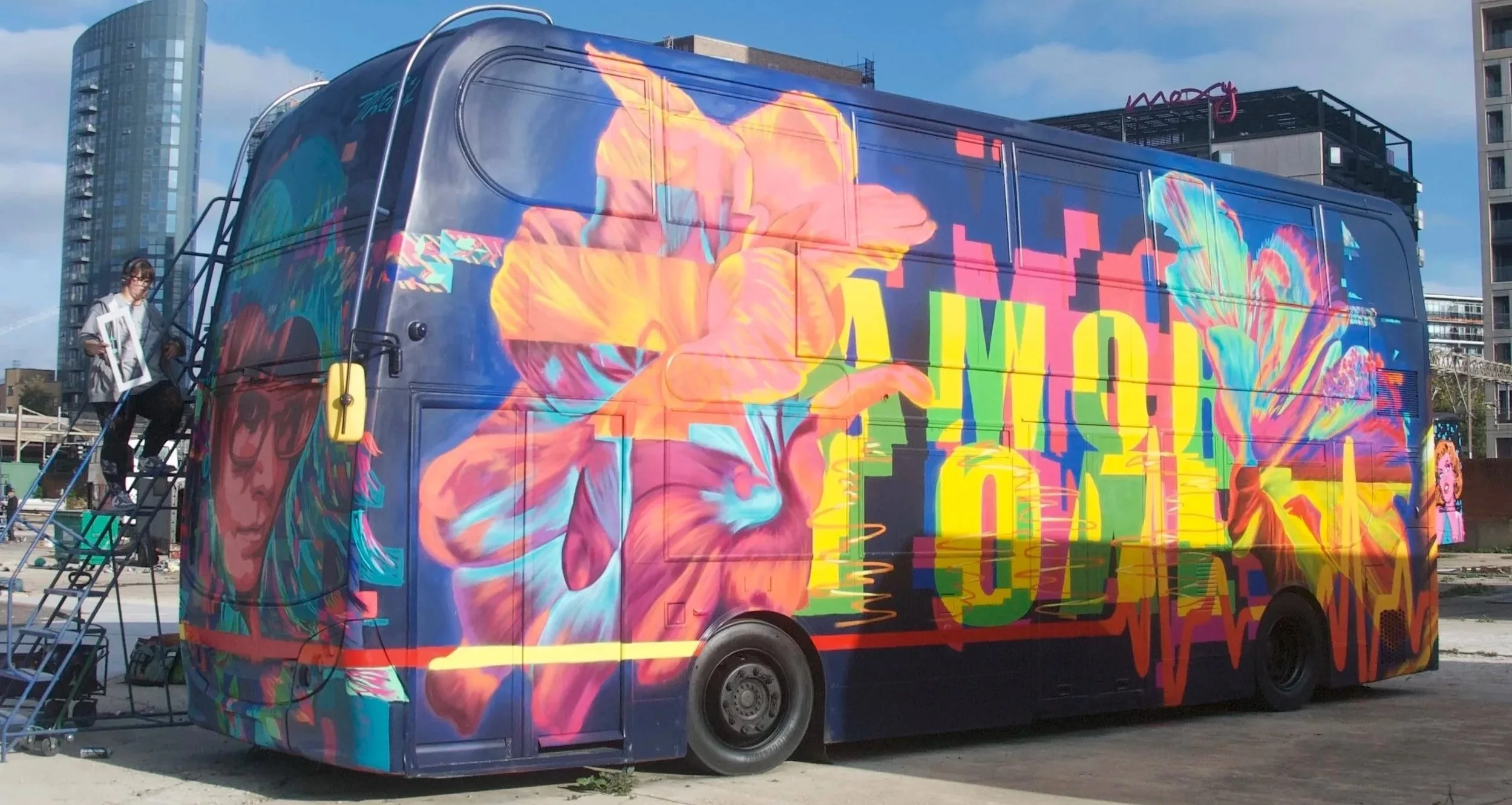 A brightly colored mural painted on a van features a woman's face with glasses, vibrant flowers, and abstract patterns, with city buildings and a person on a ladder nearby.
