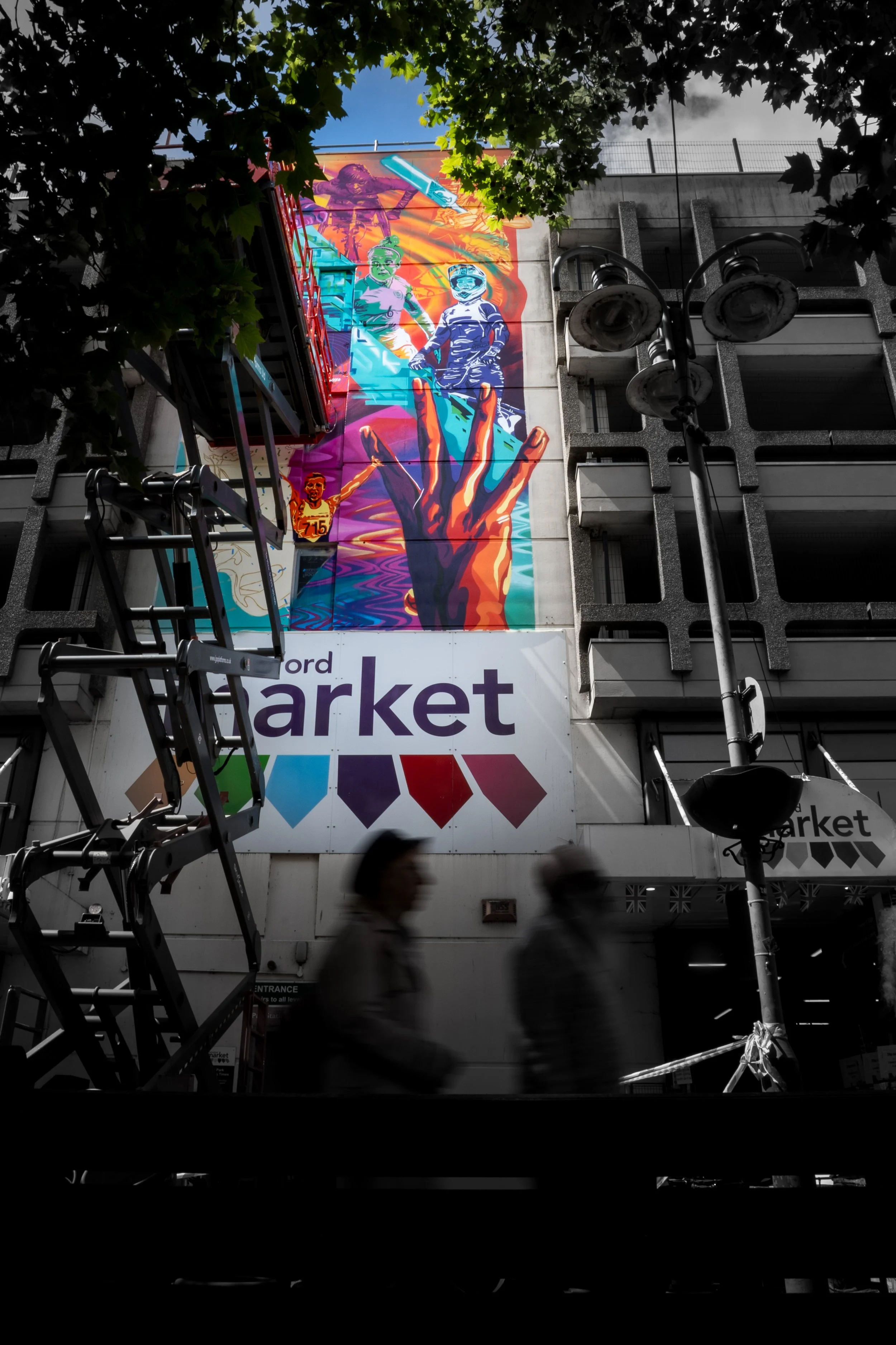 Urban scene showing a colorful mural on a building, with silhouettes of pedestrians blurred in motion, a street lamp, and some foliage partially framing the scene.
