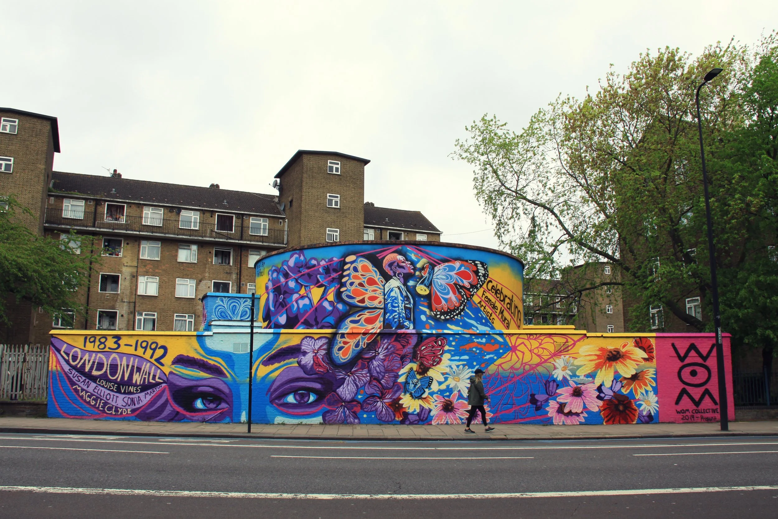 Colorful mural on an outdoor wall featuring a woman's face with butterfly and floral motifs, surrounded by trees and a brick apartment building in the background, and people walking on the sidewalk.