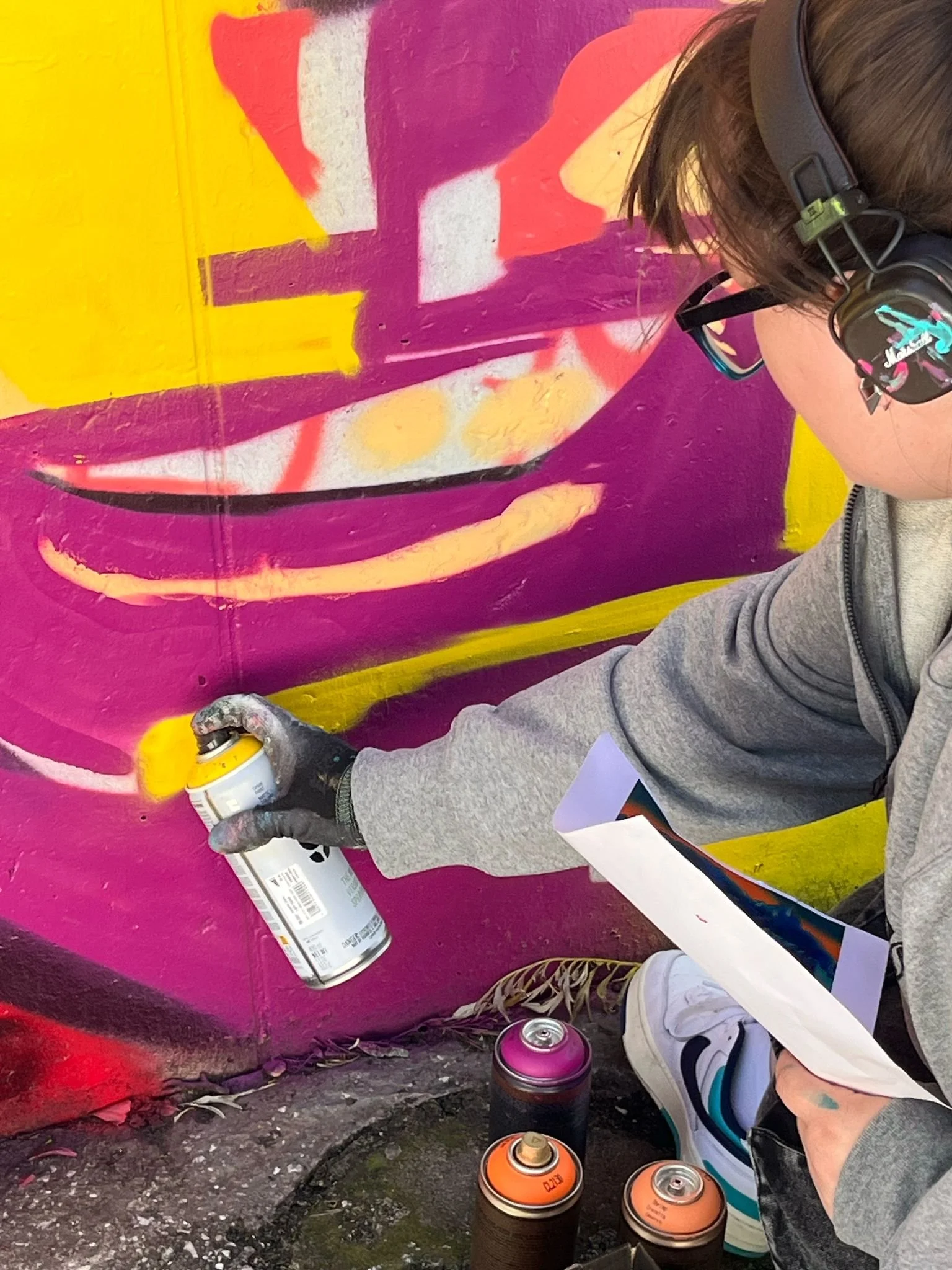 Person spray painting graffiti on a wall with a spray can in hand, wearing headphones and glasses, surrounded by multiple spray paint cans, graffiti artwork with bold yellow, purple, pink, and white colors, and a rough concrete surface.