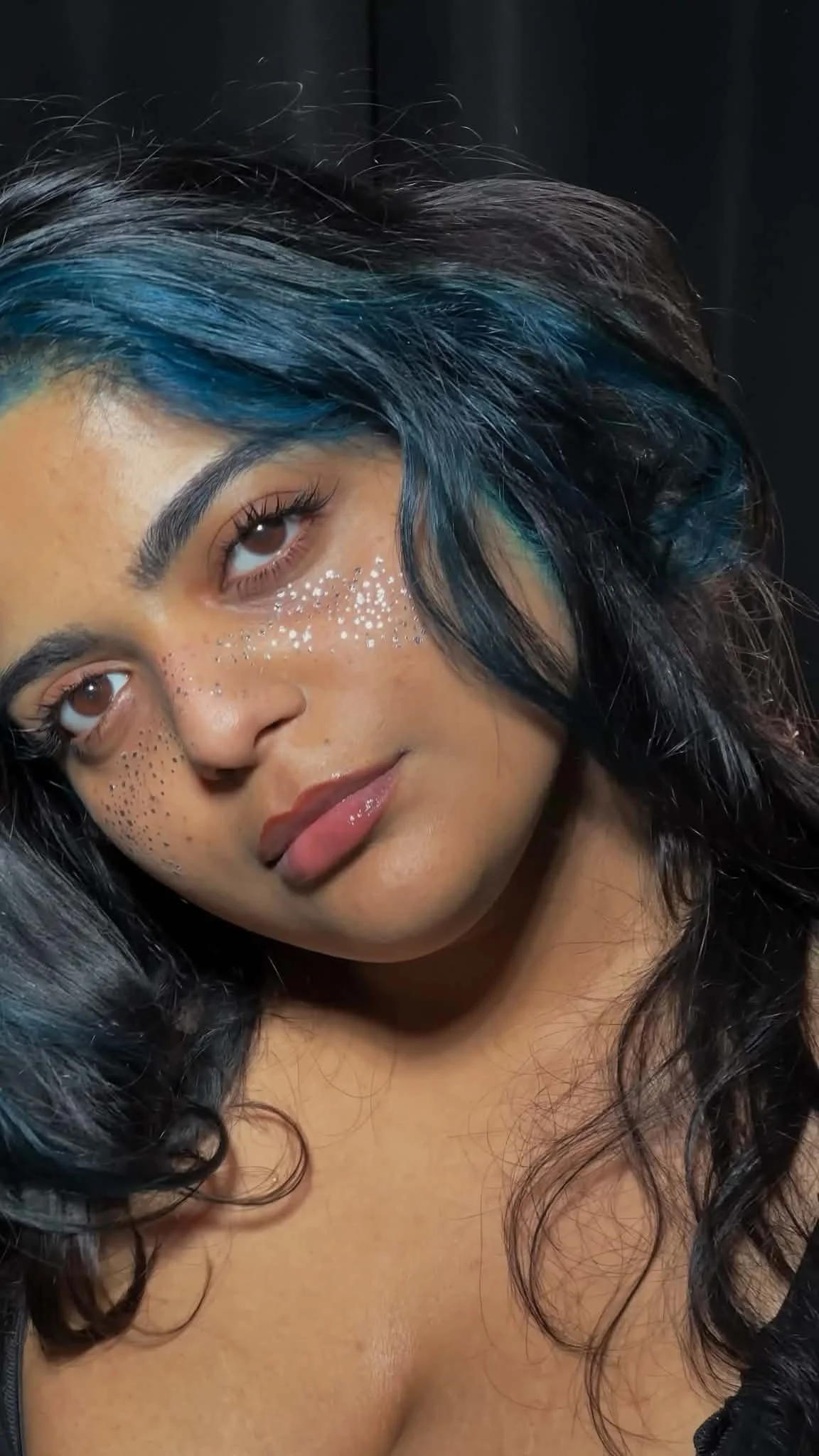 Close-up of a woman with dark curly hair with blue streaks, brown eyes, and makeup including glitter accents under her eyes.Precision artistry in every detail of this premium lash set.
