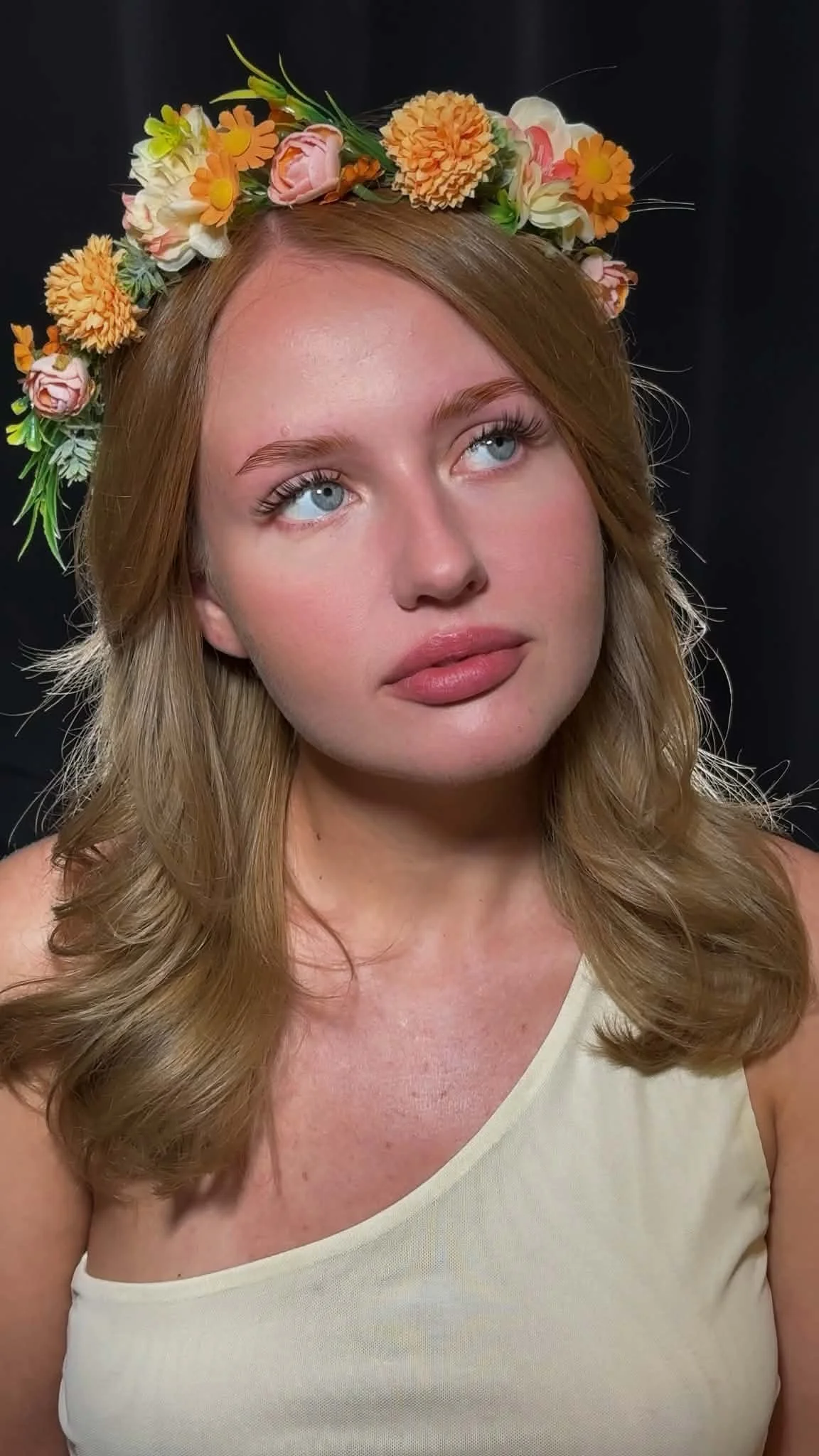 A young woman with wavy blonde hair wearing a floral crown made of peach, pink, and white flowers, and a cream-colored top, against a black background.Detailed view of luxury lash styling crafted to perfection.