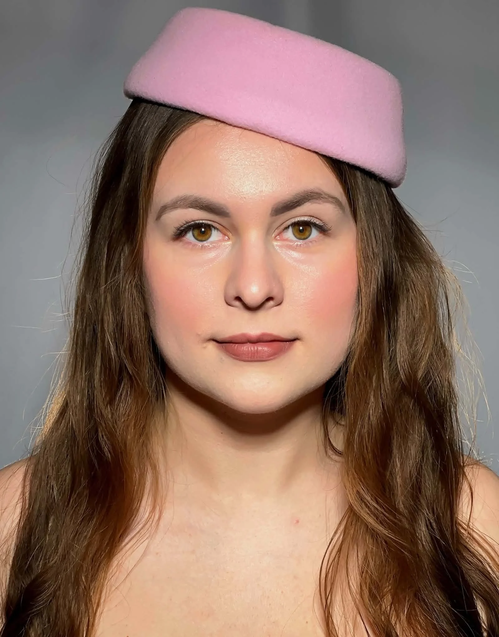 A woman with long brown hair and hazel eyes wearing a pink felt hat with a gray background.Defining elegance through expert eyelash extension artistry.