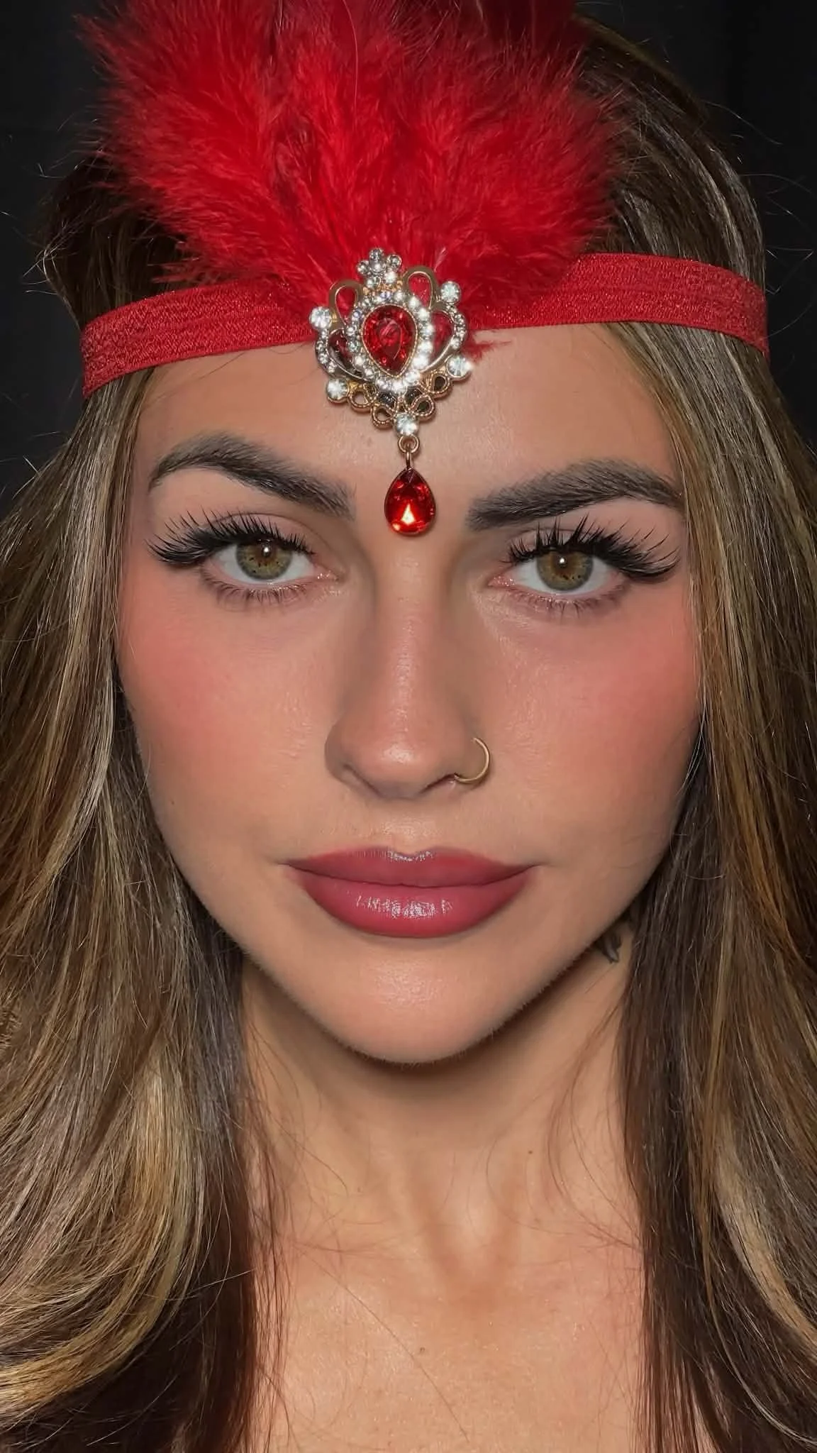 A woman with long, wavy blonde hair wearing a red headband with a feather and a decorative gemstone piece. She has light makeup, defined eyebrows, and a nose ring.Showcasing luxury eyelash enhancements and meticulous detailing by Jules Karpenko.
