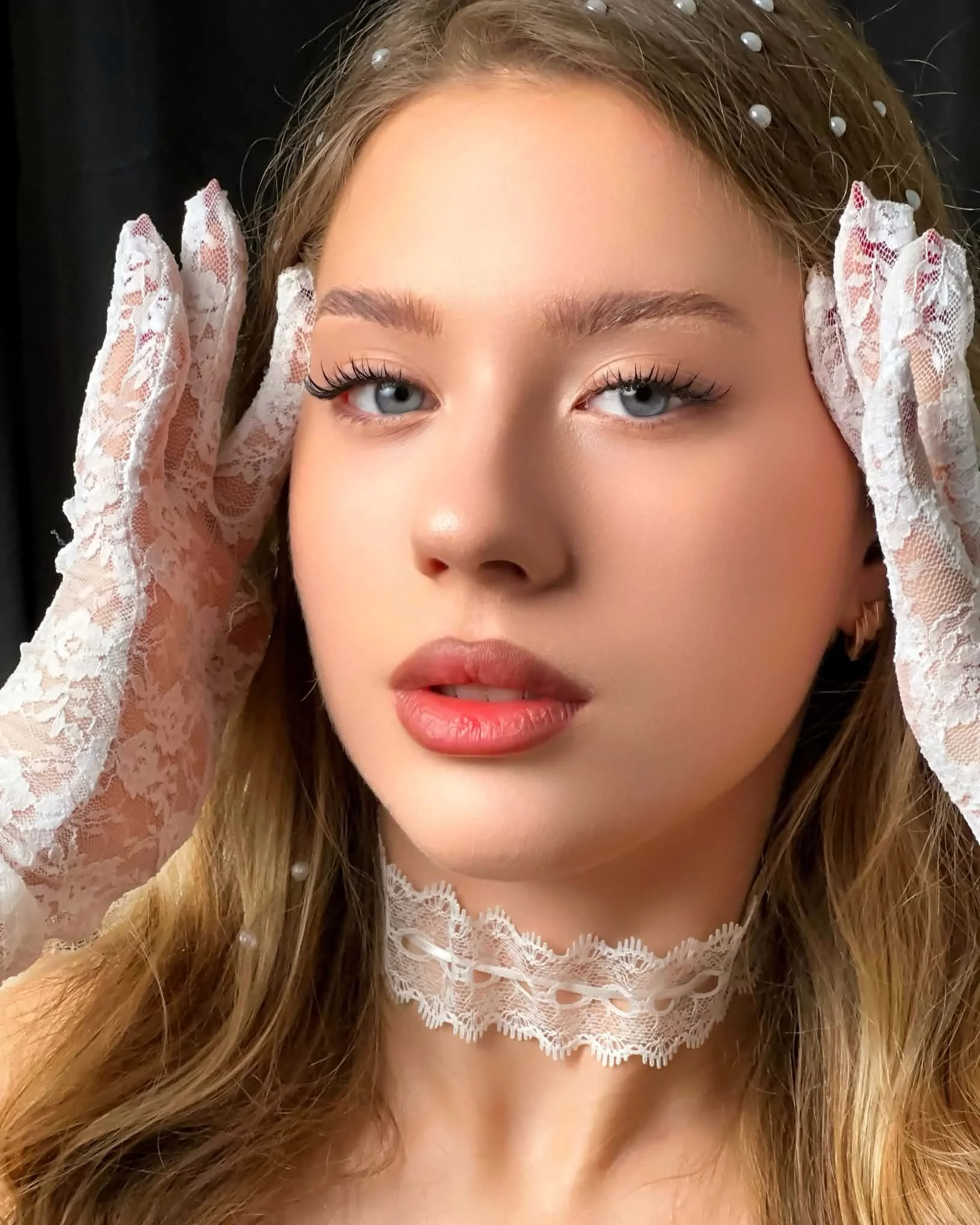 A woman with blonde hair styled in loose waves, wearing lace gloves and a lace choker, with blue eyes and natural makeup.Flawless aesthetics and superior quality lash customization.
