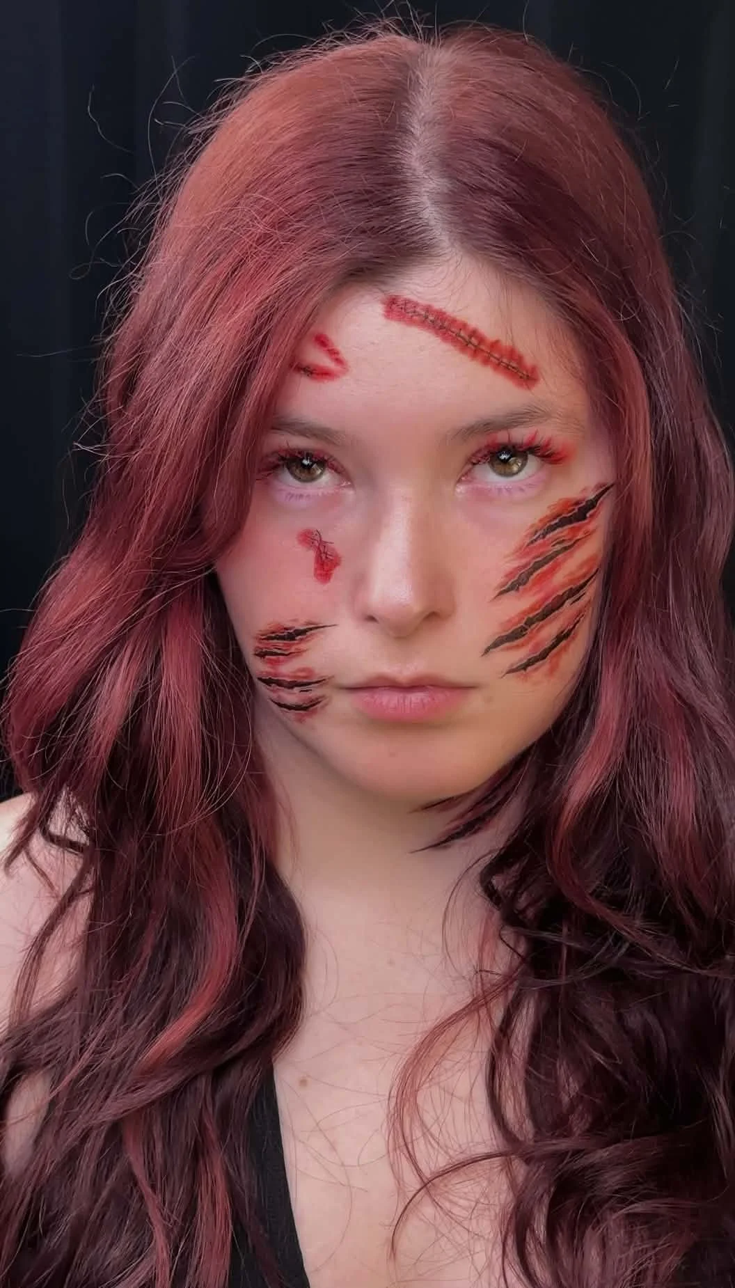 A woman with long, wavy reddish-brown hair and makeup resembling tiger stripes on her face, with false scars drawn on her forehead and cheek.This look features premium eyelash extensions and styling created by Jules Karpenko.