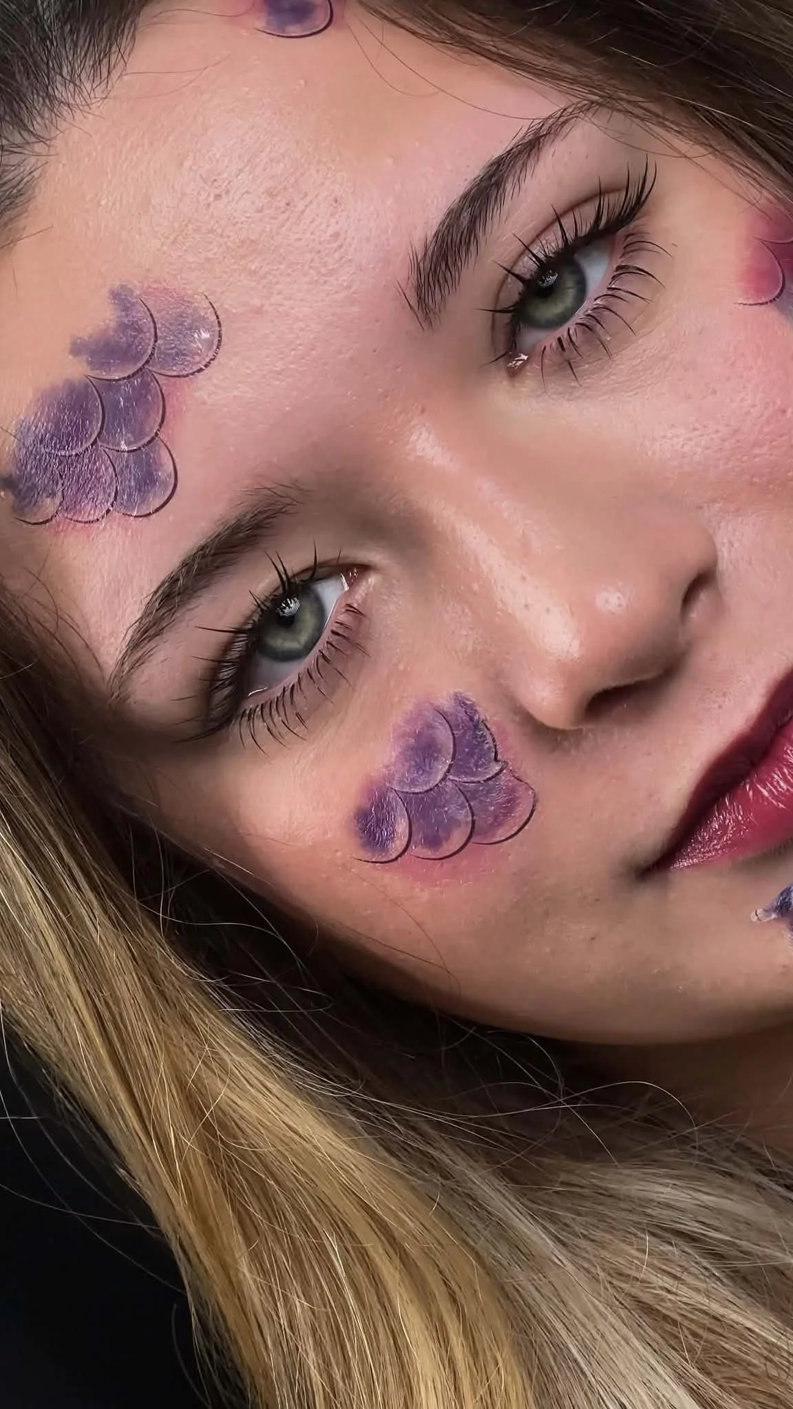 Close-up of a woman's face with artistic purple and blue lunar-themed designs on her cheeks, showcasing her clear skin, green eyes, and long lashes.Exquisite lash artistry for a sophisticated and modern beauty look by Jules Karpenko.