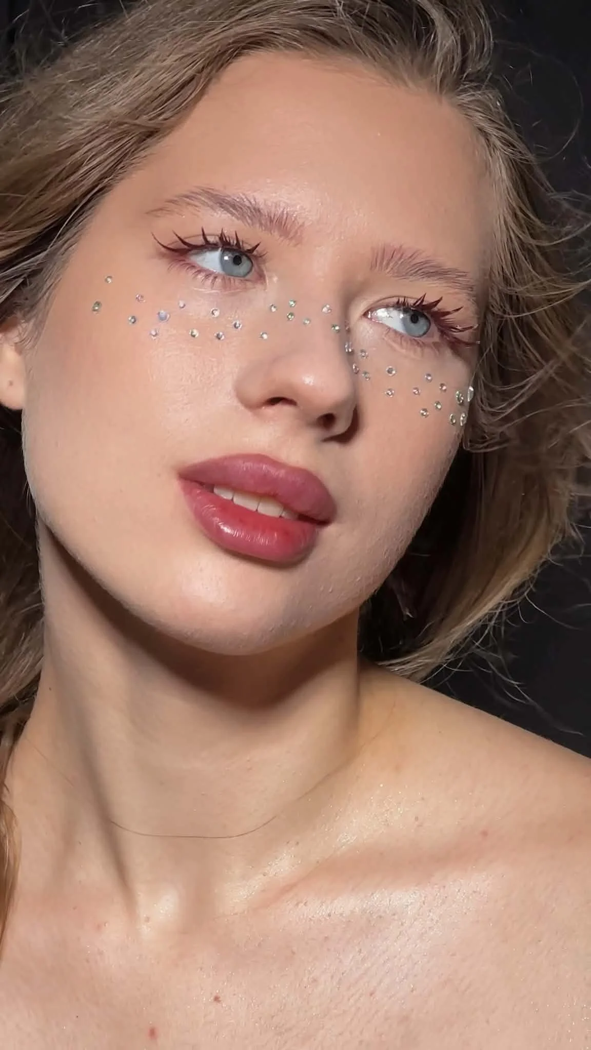 A close-up photo of a young woman with light skin, light brown hair, and blue eyes. She has small rhinestones on her cheeks and makeup including eyeliner, mascara, and natural lipstick.Portrait of a professional lash extension artist Jules Karpenko i