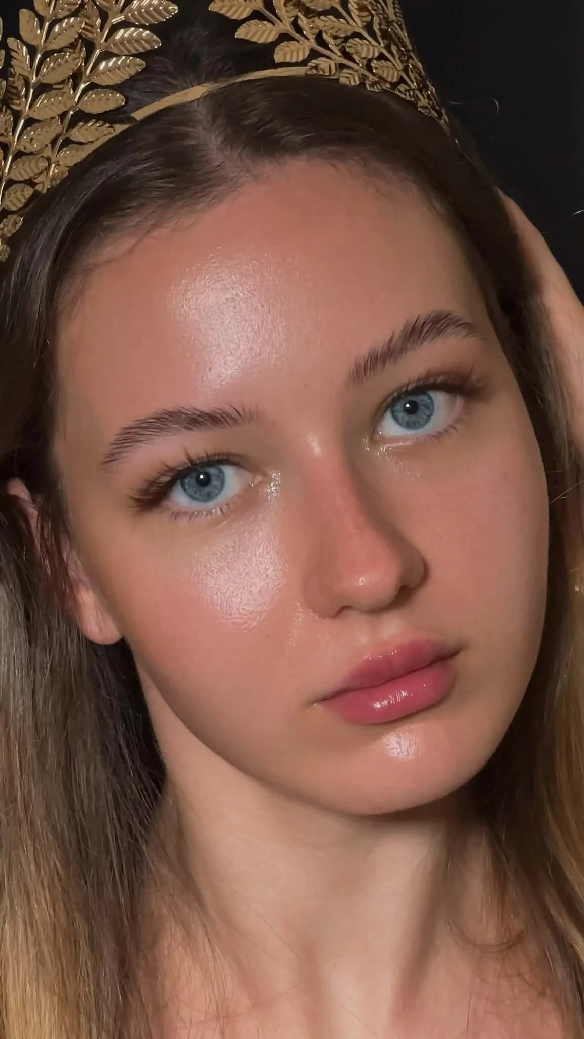 A young woman with fair skin and blue eyes, lying down with her head resting on a pillow that has a gold floral pattern. She has straight brown hair and a neutral expression.Featuring handcrafted eyelash artistry tailored by Jules Karpenko.