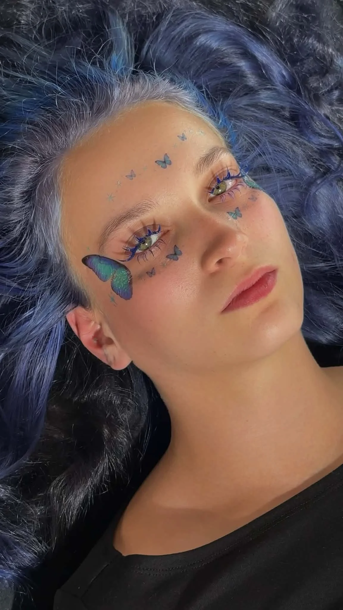 A woman with blue-tinted hair lying down, featuring butterfly-shaped face tattoos around her eyes and forehead.Highlighting the artistry of premium lash extension application by Jules Karpenko.