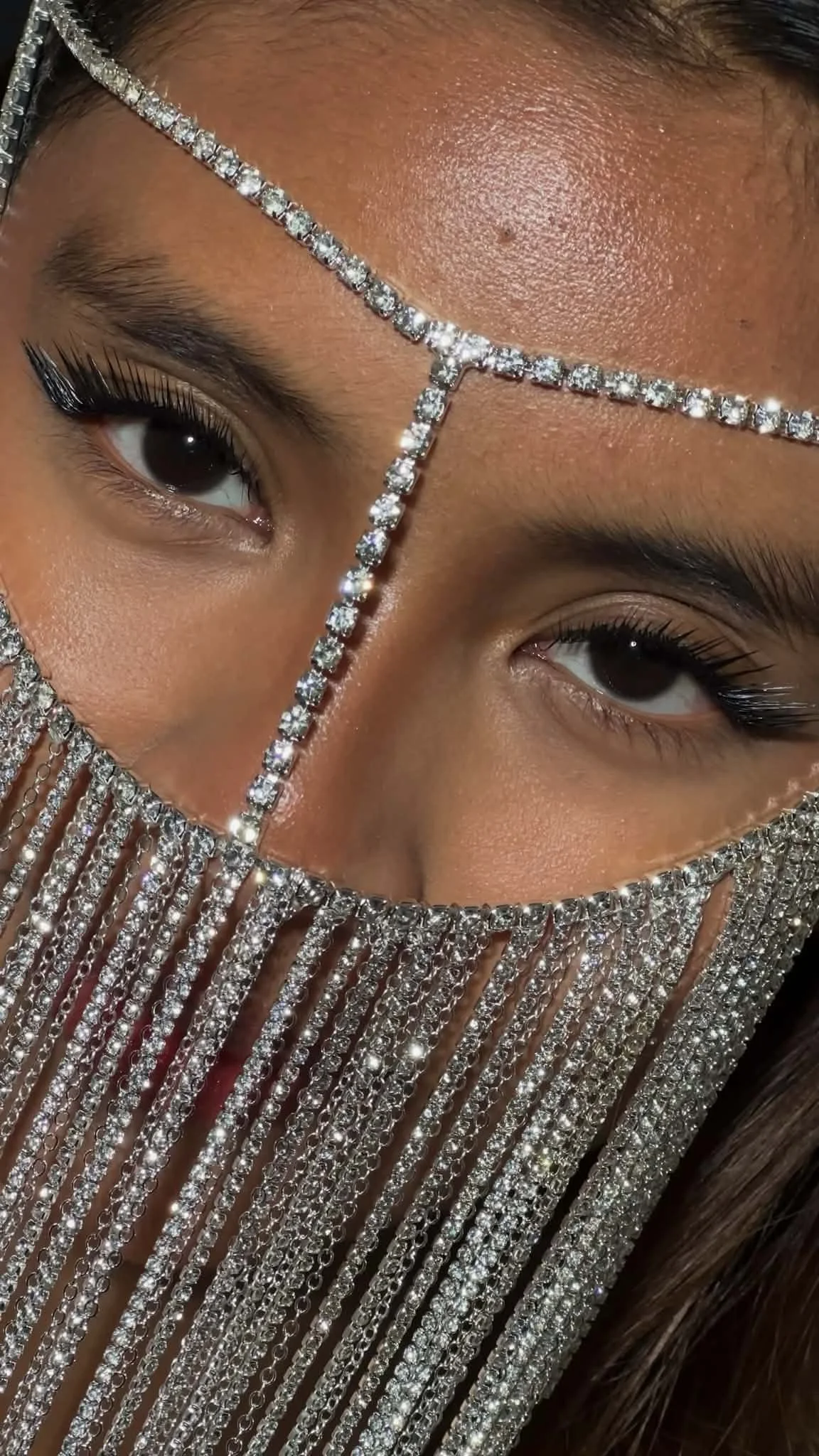 Close-up of a person's face adorned with diamond jewelry, a headpiece, and dangling chains covering part of their face, highlighting their eyes and skin.Showcasing bespoke lash elegance and superior lash design techniques.
