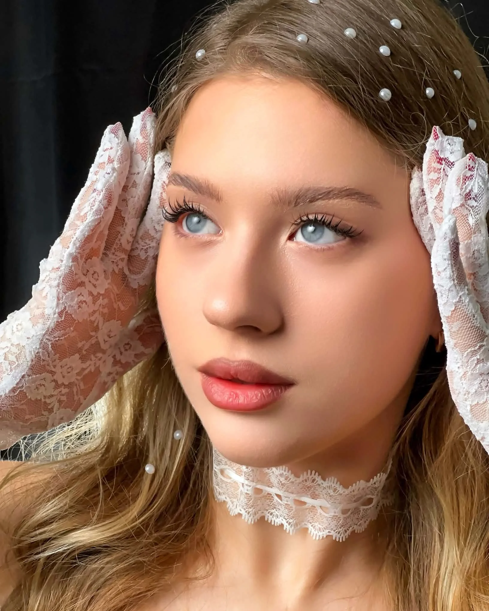 Close-up of a young woman with blue eyes, wearing lace gloves and a string of pearls in her hair, gazing slightly upward.Editorial fashion combined with luxury lash artistry.