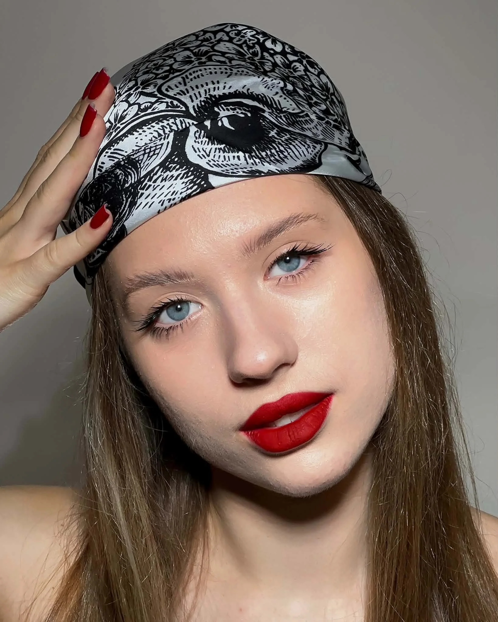 Close-up of a young woman with blue eyes and red lipstick wearing a black and white patterned headscarf, with her hand touching the side of her head.A stunning display of volume lash extensions for an editorial look by Jules Karpenko.