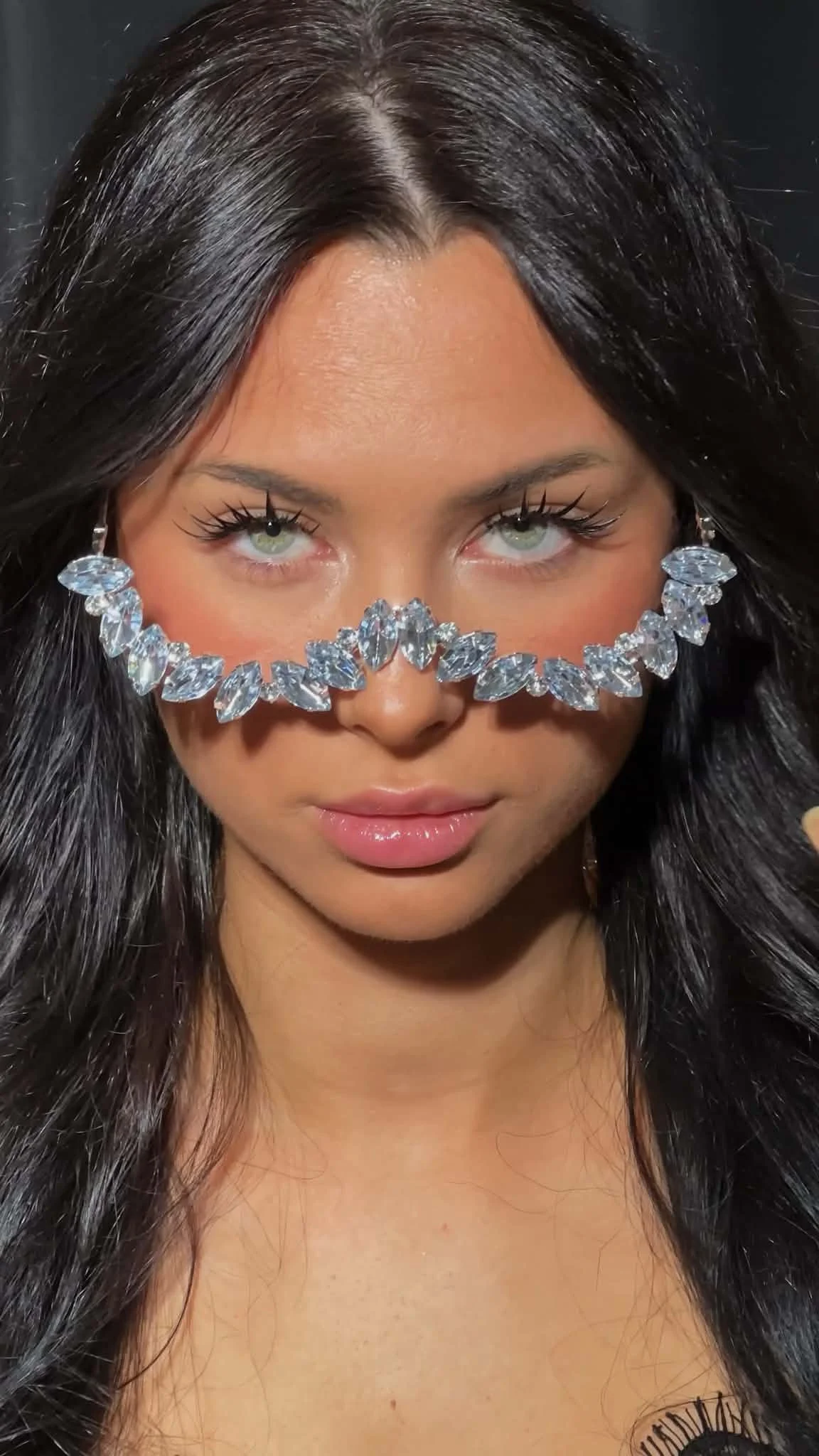A woman with dark hair and green eyes wearing a face mask adorned with clear, gem-like stones.Displaying precision lash styling and elegant beauty design by Jules Karpenko.