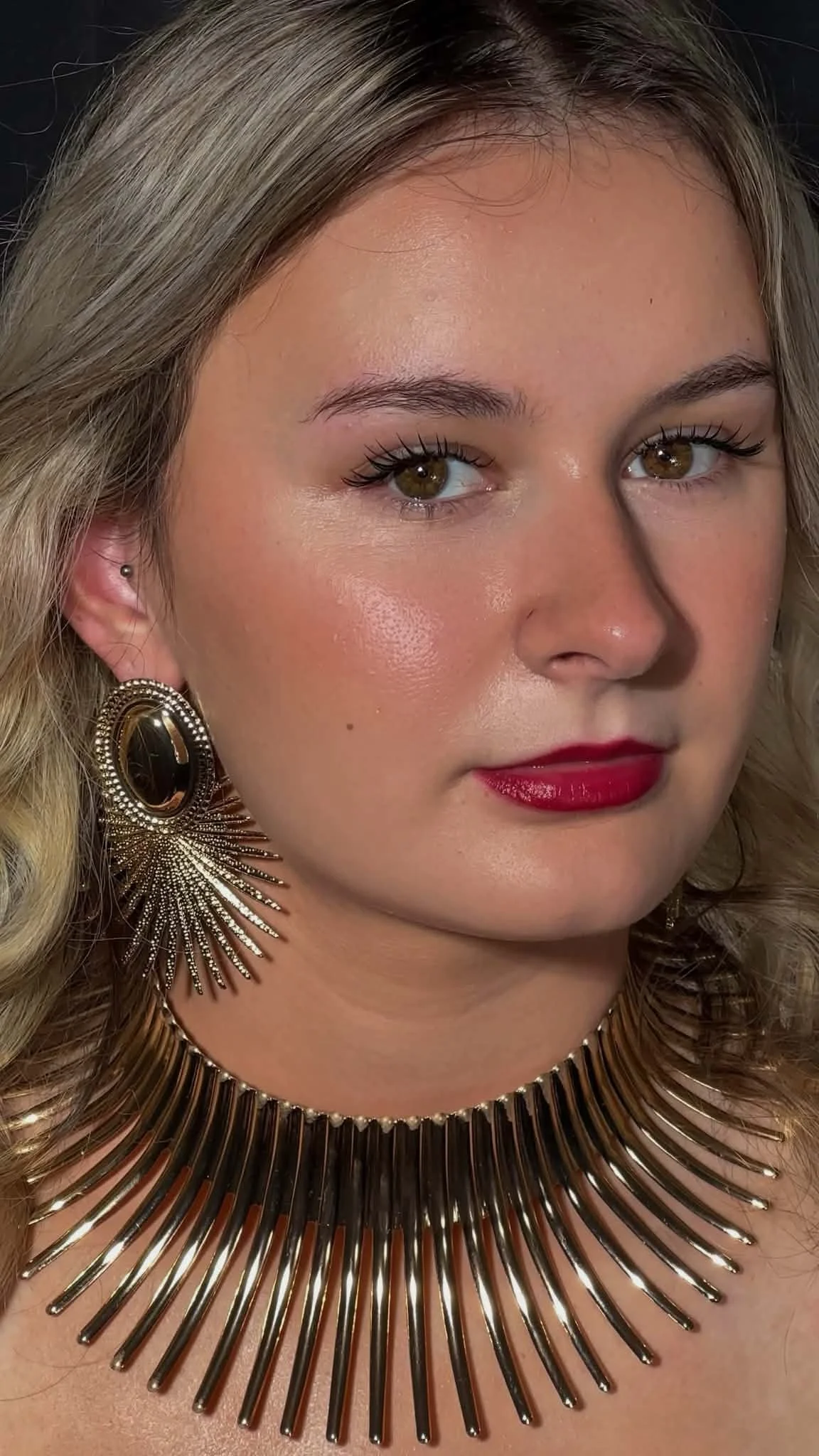 A woman wearing large, ornate gold earrings and a matching gold statement necklace.Flawless eyelash extension application ensuring a natural yet dramatic effect by Jules Karpenko.
