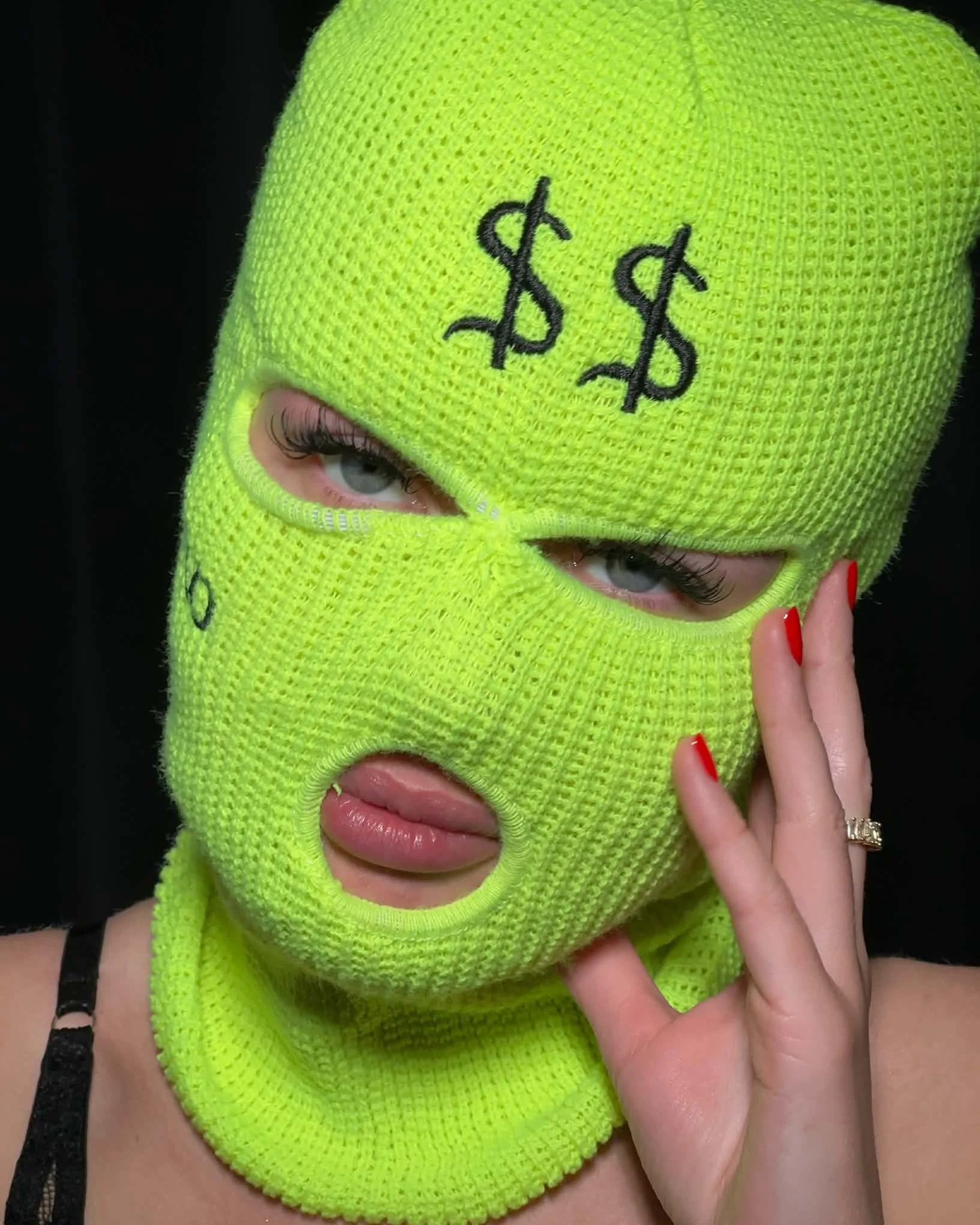 Person wearing a bright yellow knit balaclava with black dollar signs embroidered on it, revealing only one eye and hand with red nail polish.A masterpiece in eyelash design featuring custom volume and style.