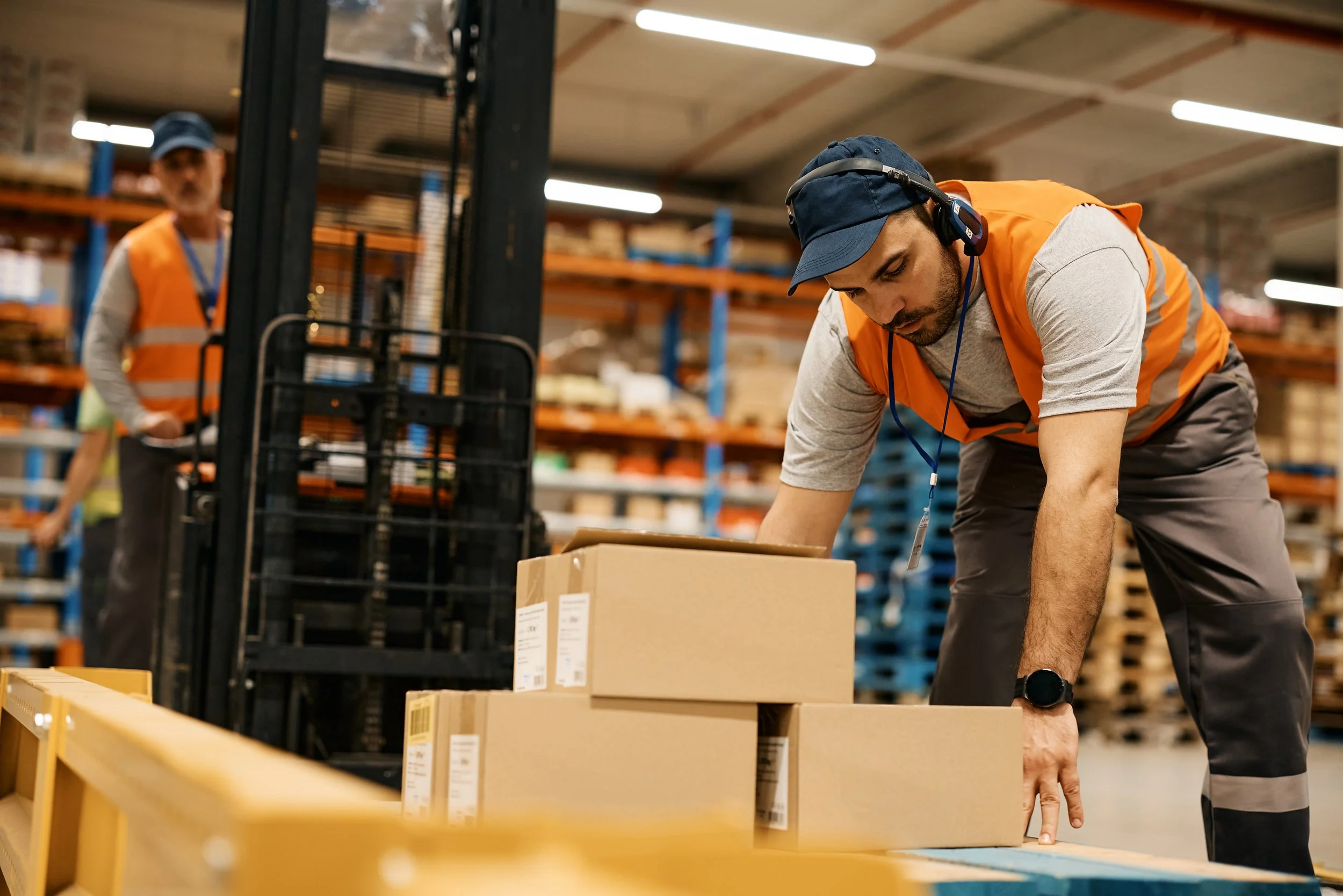 A warehouse worker wearing a gray t-shirt, orange safety vest, blue cap, and headphones is bending over and pointing at a cardboard box on a yellow table, with another worker in the background. Shelves with various boxes and goods are visible in the warehouse.