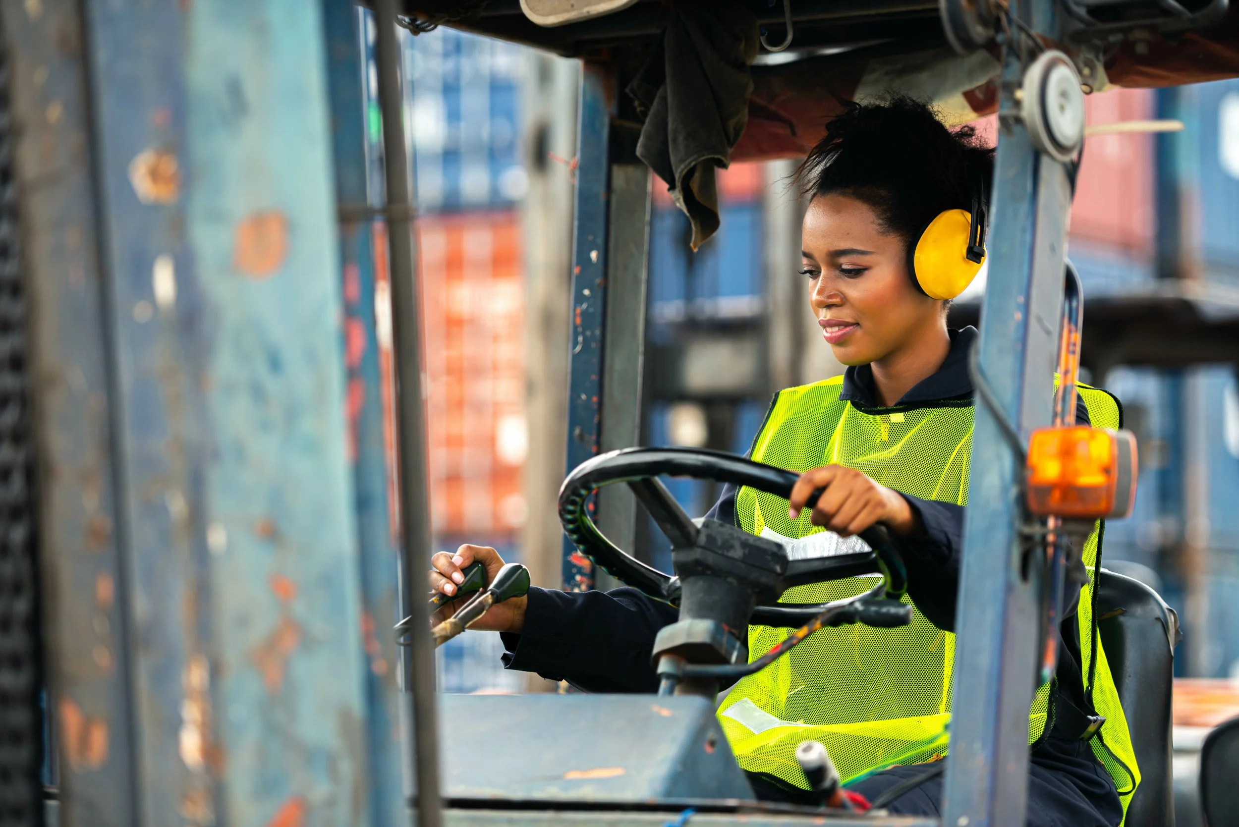 A woman wearing a yellow safety ear protector and a neon yellow reflective safety vest is operating a forklift in an industrial setting.