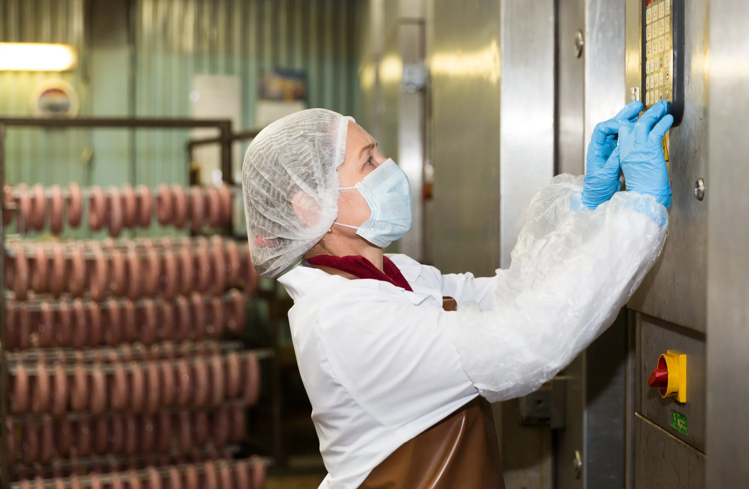 A woman wearing a hairnet, face mask, and gloves operating an industrial machine in a food processing facility, with rows of processed meat products in the background.