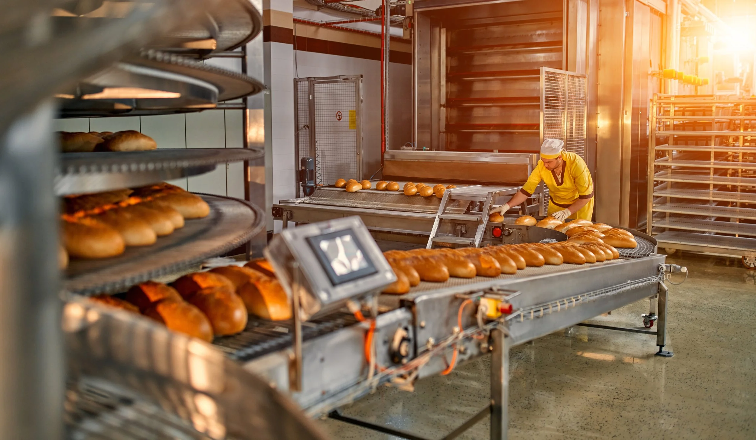 A bakery worker wearing a yellow uniform and white cap is inspecting freshly baked bread on a conveyor belt in a large industrial bakery. Several trays of bread are visible on metal racks in the foreground, with sunlight streaming in from the right side of the image.