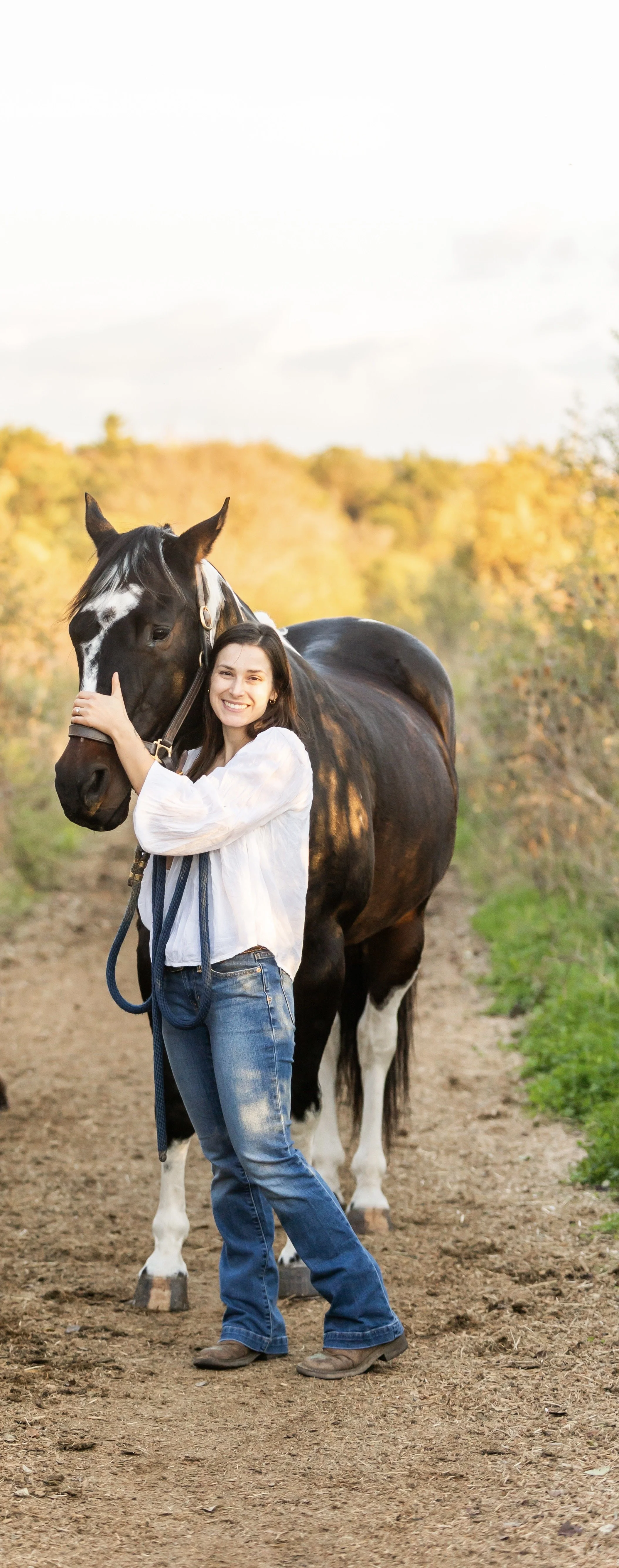 A woman in a white blouse and jeans standing on a dirt path, smiling and hugging a black and white horse with a background of trees with autumn foliage.