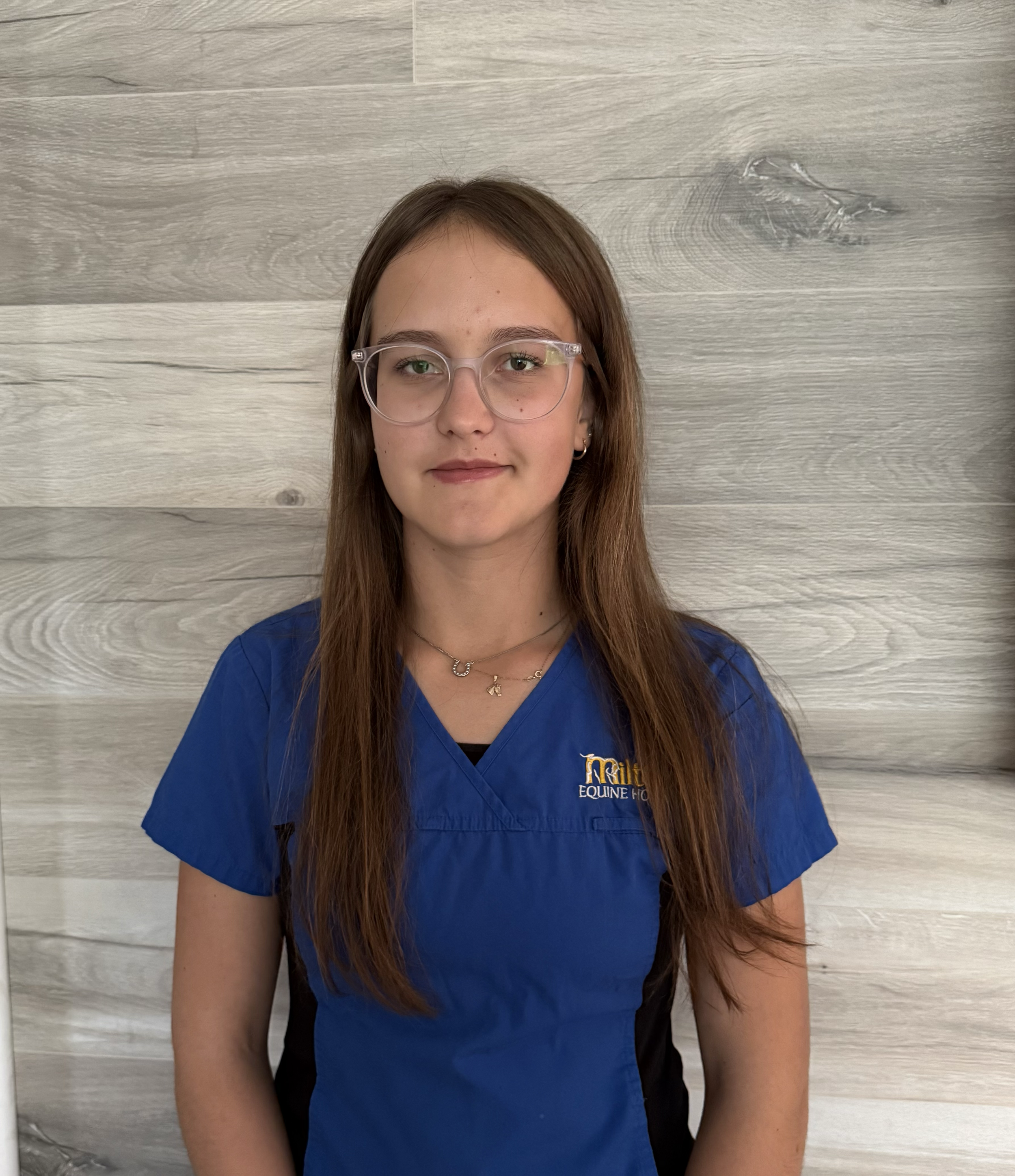 A young woman with long brown hair wearing clear glasses and a blue uniform top, standing in front of a light wooden wall.
