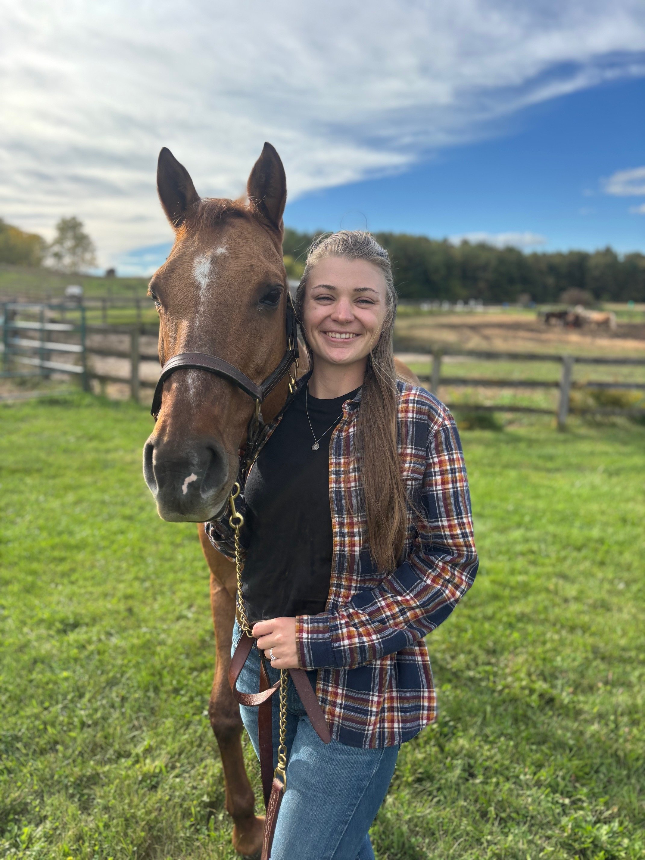 A young woman with long hair smiling stands next to a brown horse on a grassy field with a fence, trees, and a partly cloudy sky in the background.