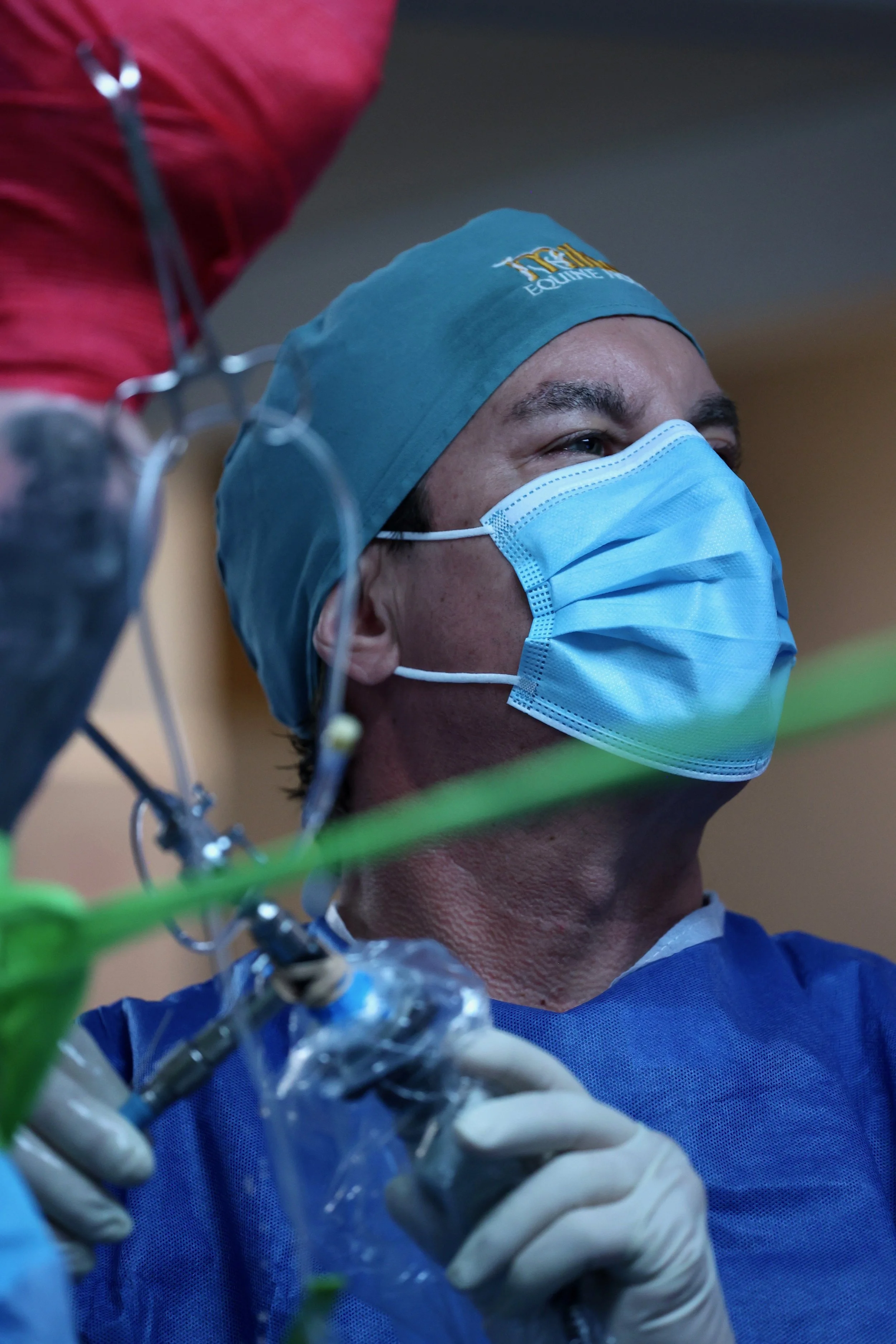A male medical professional wearing a surgical mask, blue scrubs, and a surgical cap, working with medical equipment, possibly in a hospital or operating room.