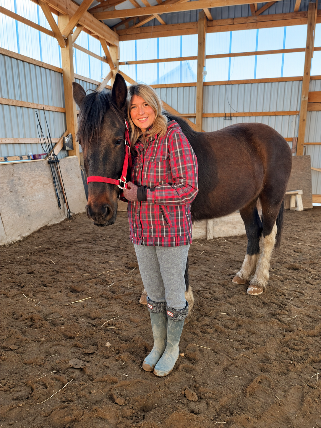 A woman standing in a barn, holding the head of a brown and black horse with a red halter, both smiling at the camera. The barn has wooden beams and a dirt floor.