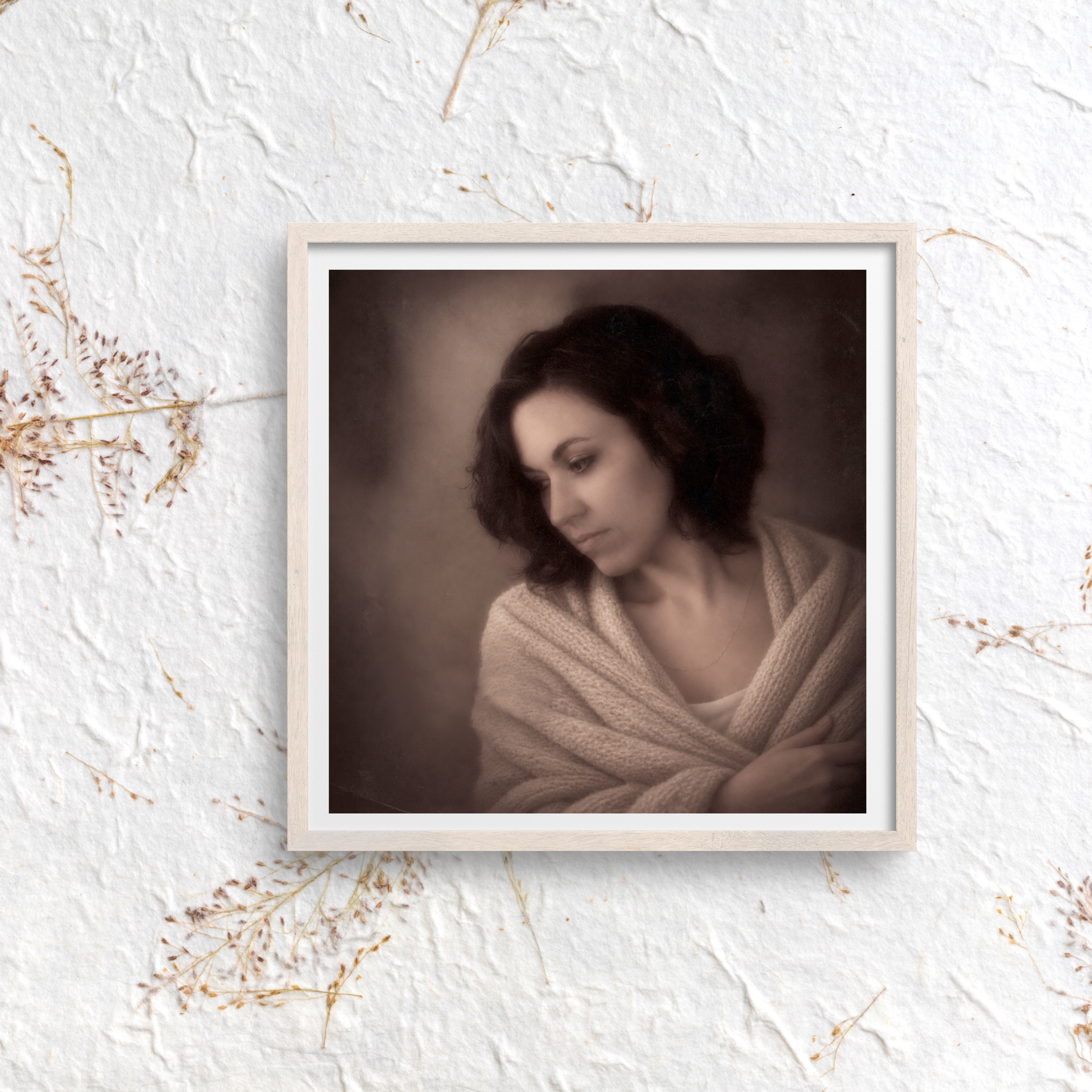 Close-up of a framed portrait photograph of a woman with dark, curly hair, set against a textured white wall with dried plants on the side.