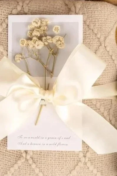 A small bouquet of white dried flowers wrapped with a white satin ribbon, placed on a white card with text, resting on a beige textured fabric surface.