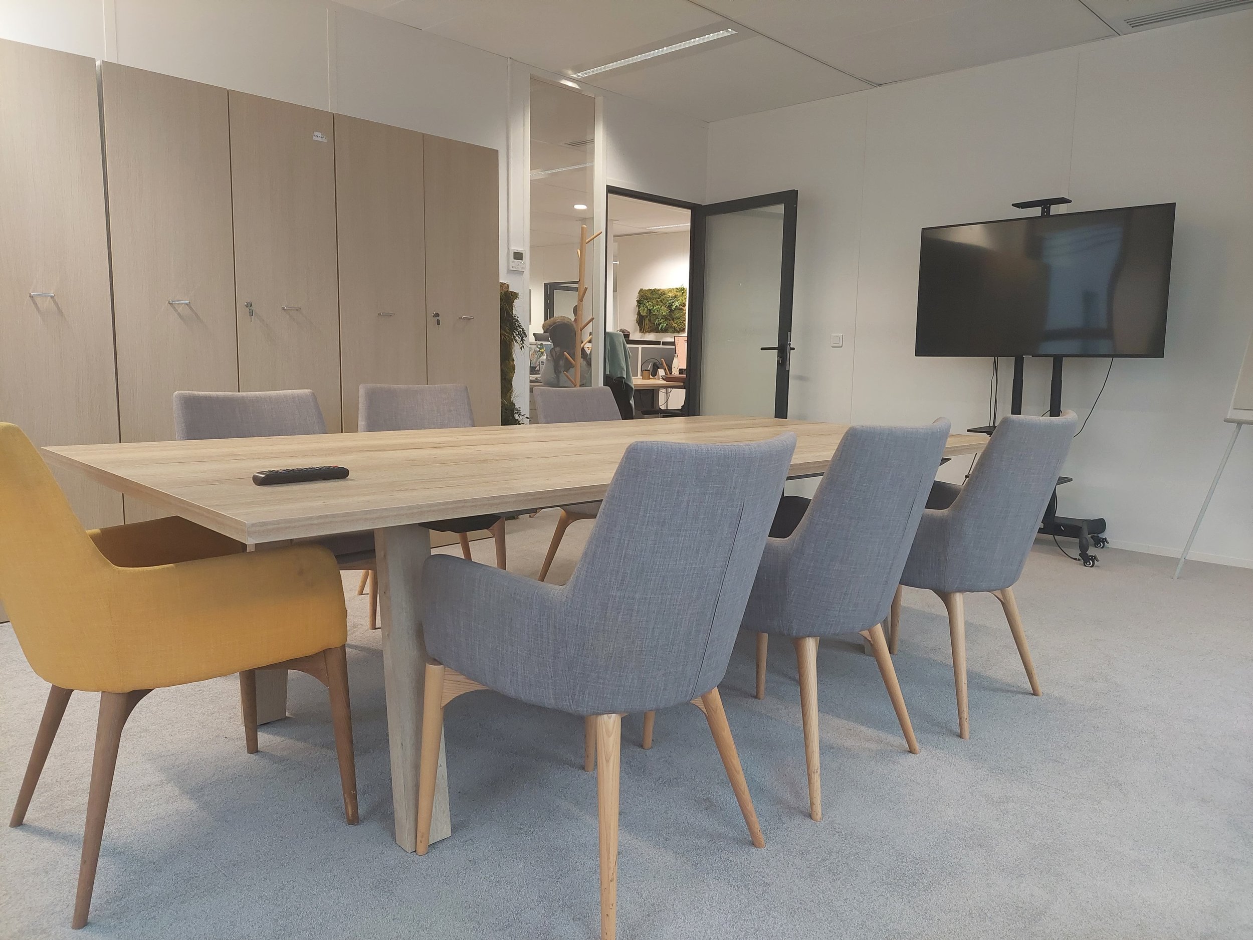 Une salle de réunion avec une table en bois, plusieurs chaises en tissu gris, un grand écran noir sur un support, un mur blanc, des armoires en bois, une porte ouverte menant à un espace de bureau avec une personne à son bureau, et des plantes décoratives.