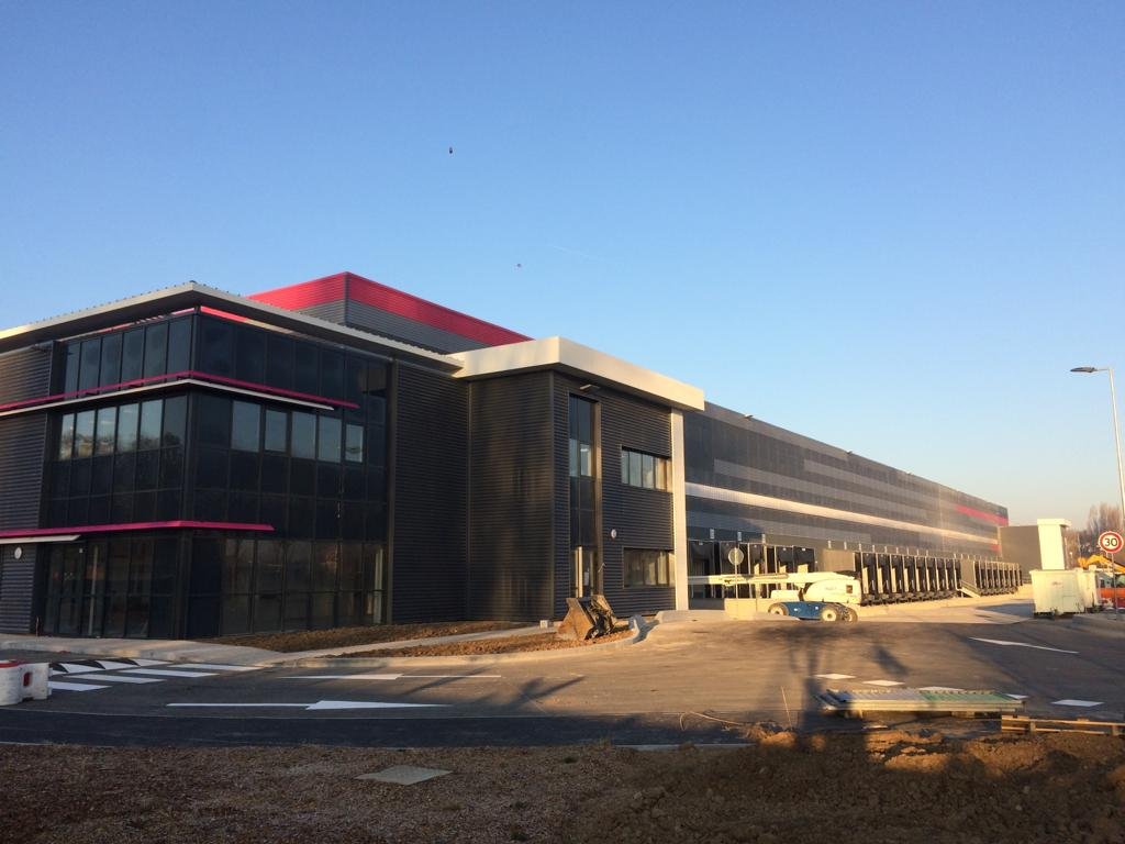 Un grand bâtiment moderne avec des murs en verre noir et des détails roses et blancs, construction en cours avec des véhicules de chantier et des matériaux.