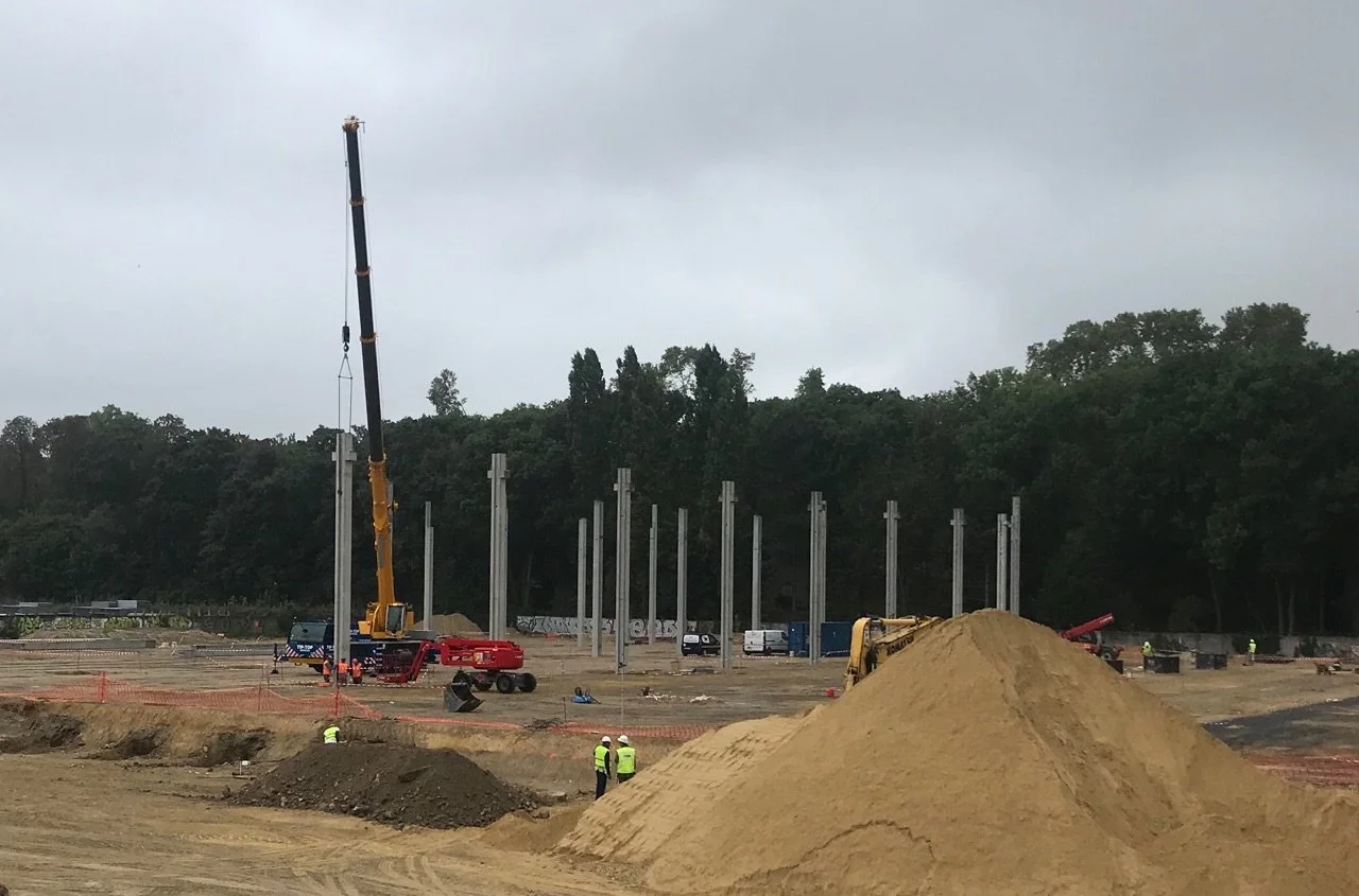 Chantier de construction avec un grue en action, des travailleurs en gilet de sécurité, une pile de terre, et des piliers en acier en arrière-plan.