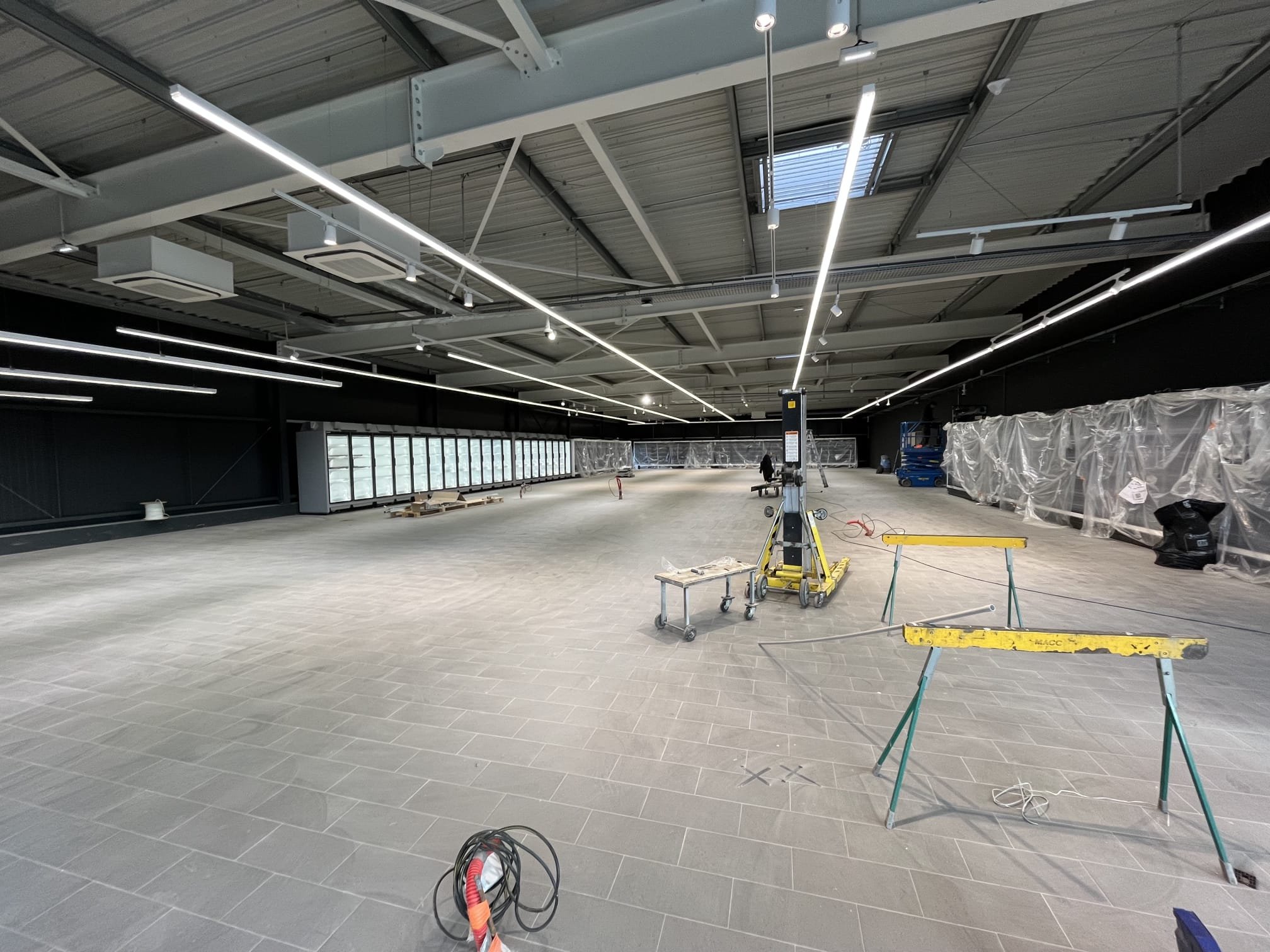 Grand espace intérieur en cours de construction ou rénovation avec plafonds hauts, éclairage suspendu LED, revêtement de sol en carrelage gris, murs noirs, matériaux et outils de construction. Il y a des structures métalliques, des fils électriques, et du matériel de chantier, avec un panneau de contrôle ou de signalisation au centre.