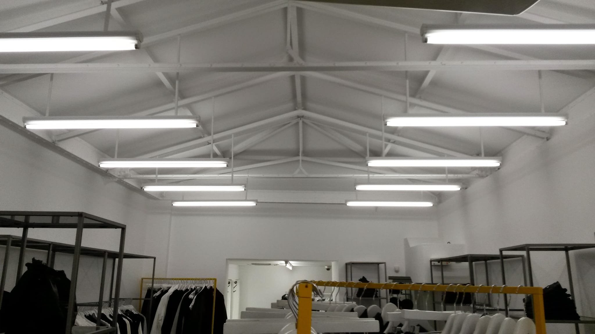Intérieur d'un entrepôt ou d'une boutique avec des étagères métalliques et des racks contenant des vêtements. Le plafond est en poutres métalliques peintes en blanc avec de longues lumières fluorescentes.