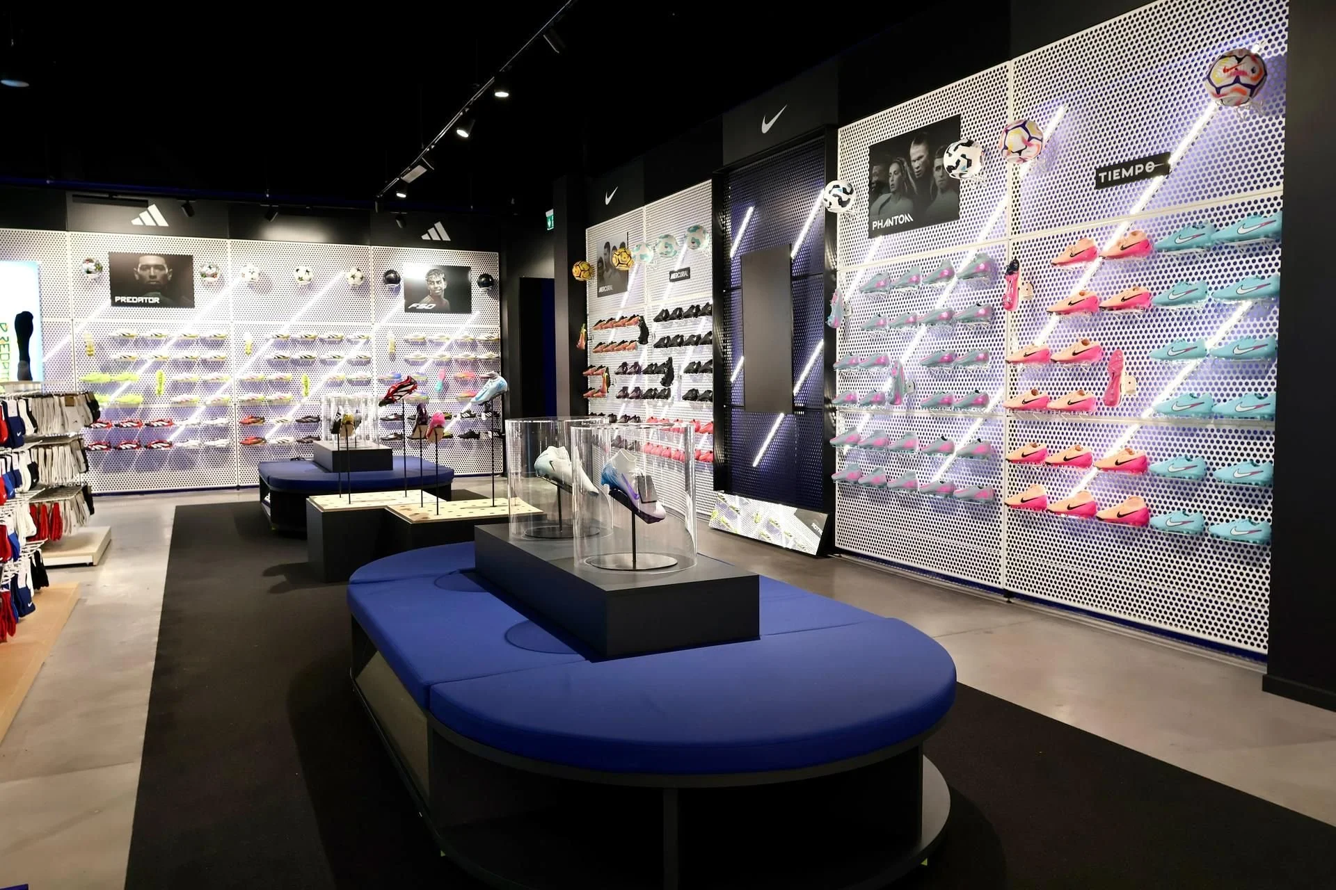 Intérieur d'un magasin de sport avec des chaussures de football colorées sur un mur à droite, des ballons de football suspendus et des affiches de joueurs célèbres comme Cristiano Ronaldo et Neymar.