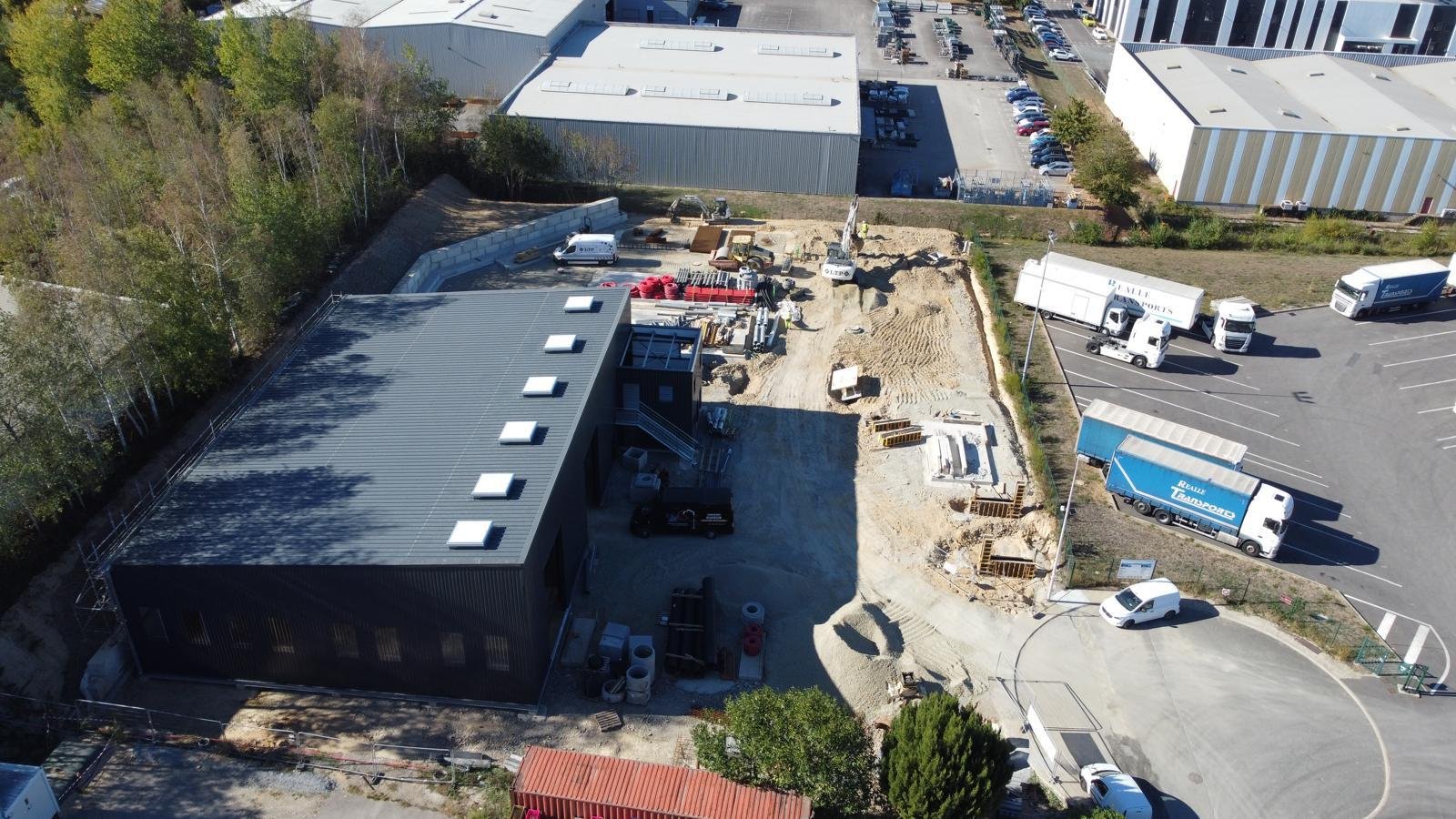 Chantier de construction avec un bâtiment neuf, véhicules de chantier, un coffrage, et un parking avec des camions
