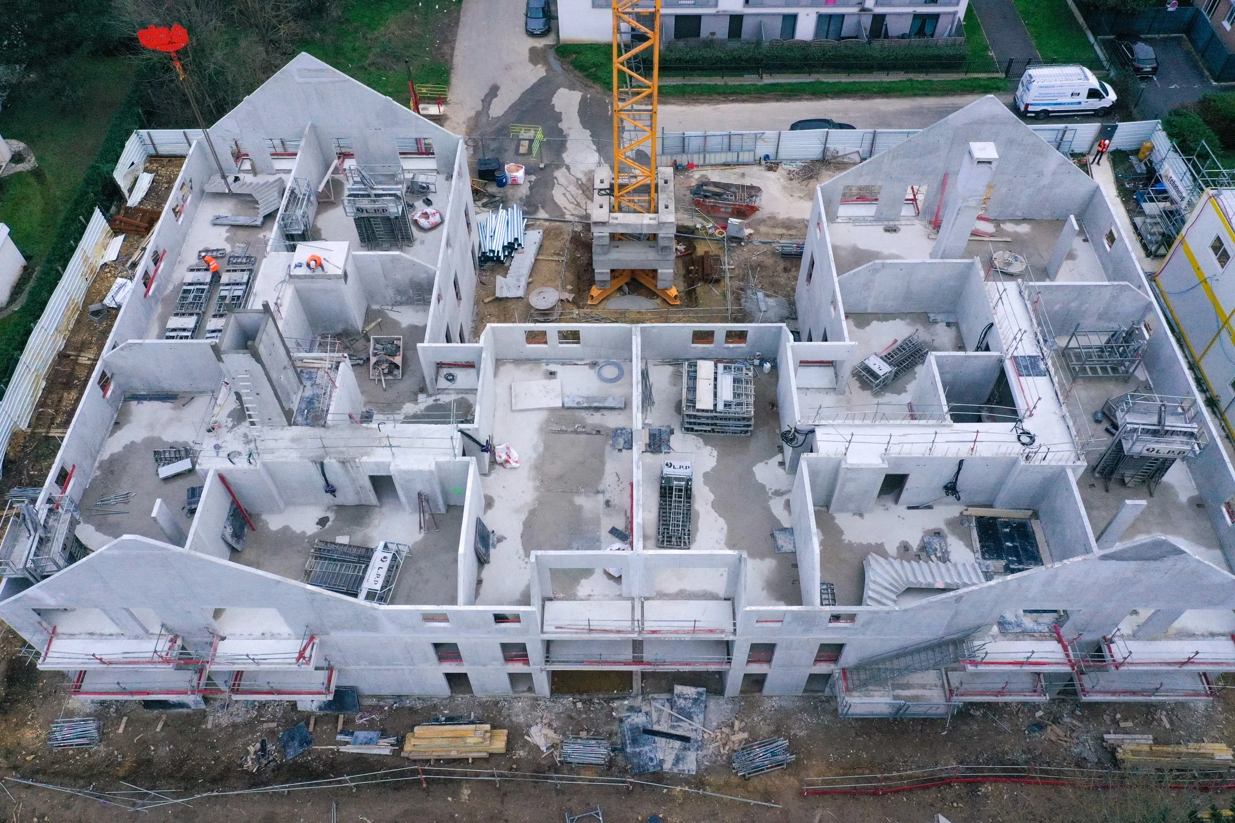 Vues aériennes d'un bâtiment en construction avec une grue orange au centre, murs en béton, matériaux de construction et véhicules à proximité.