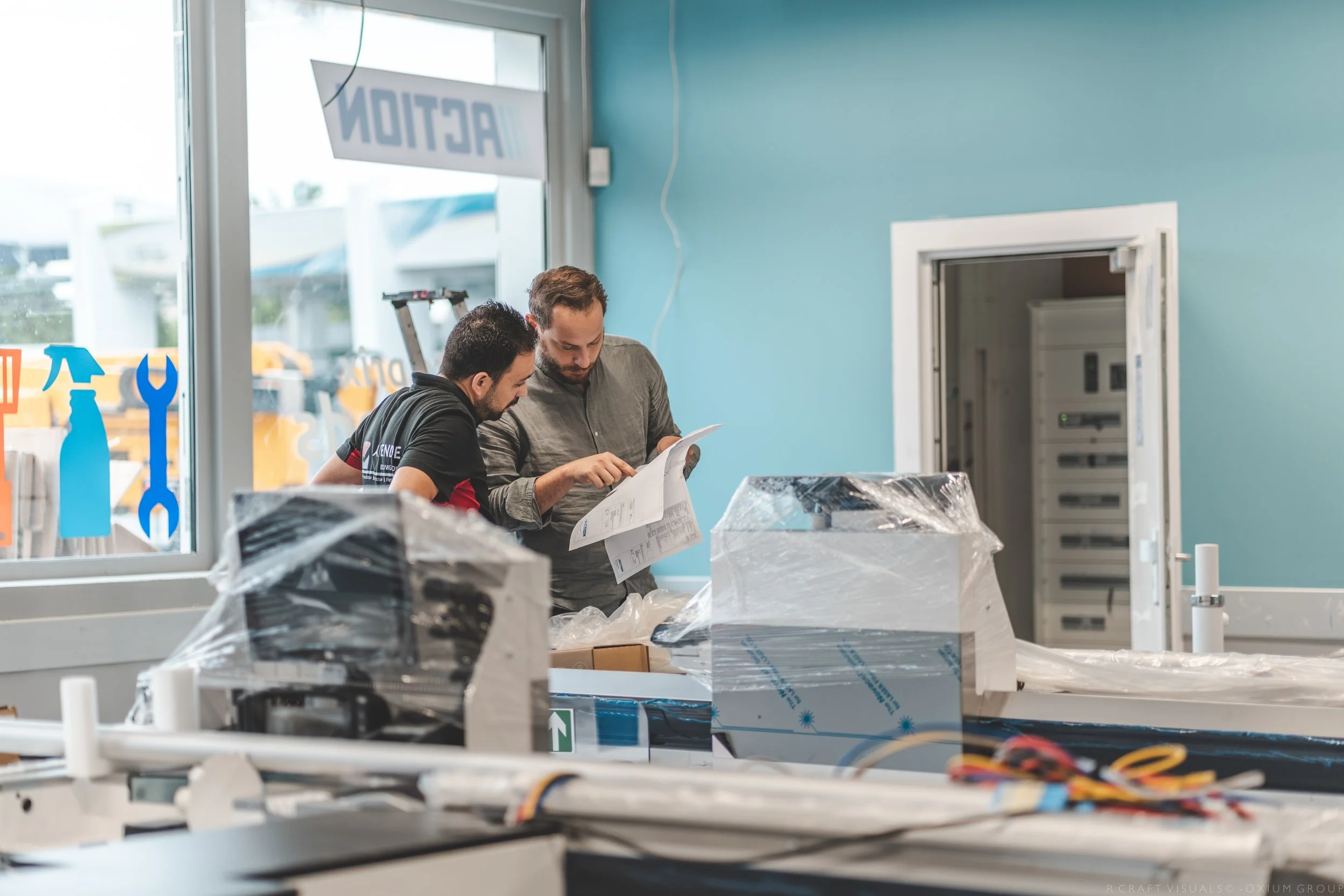 Deux hommes regardent un document dans un atelier ou un magasin, entourés de matériel et d'emballage. Un panneau indique 'ACTION' et il y a des fenêtres avec des symboles de bricolage.