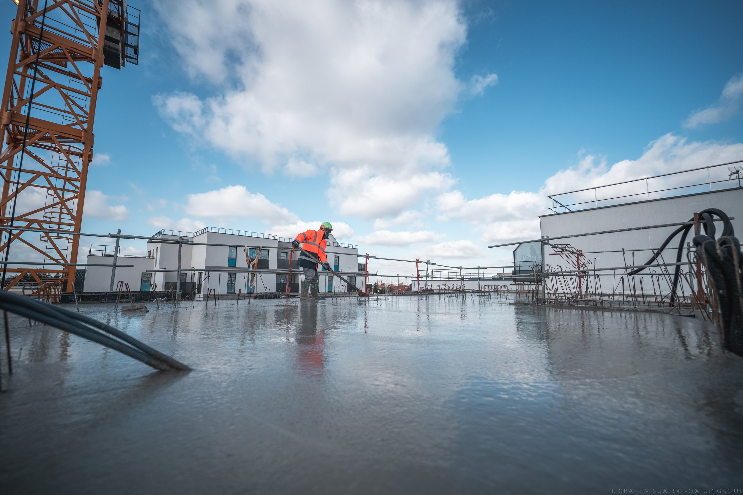Chantier de construction avec un ouvrier élégamment habillé en veston à rayures, casque de sécurité vert et gilet de sécurité orange, travaillant sur une surface en béton humide, avec des bâtiments modernes en arrière-plan et un ciel bleu avec quelques nuages.