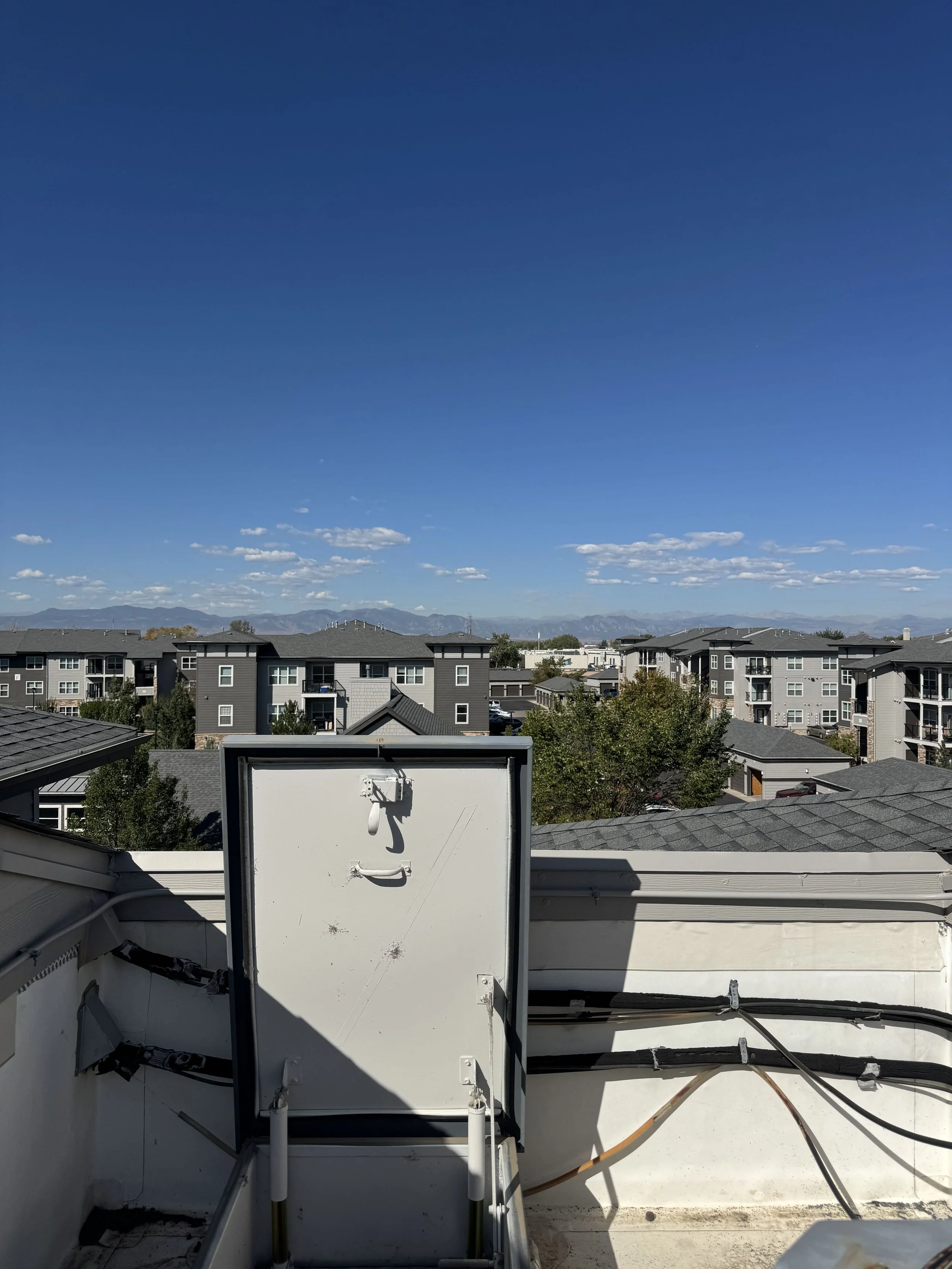 View of rooftops in a neighborhood with mountains in the distance under a partly cloudy sky, taken from a balcony with HVAC equipment and electrical wires in the foreground.