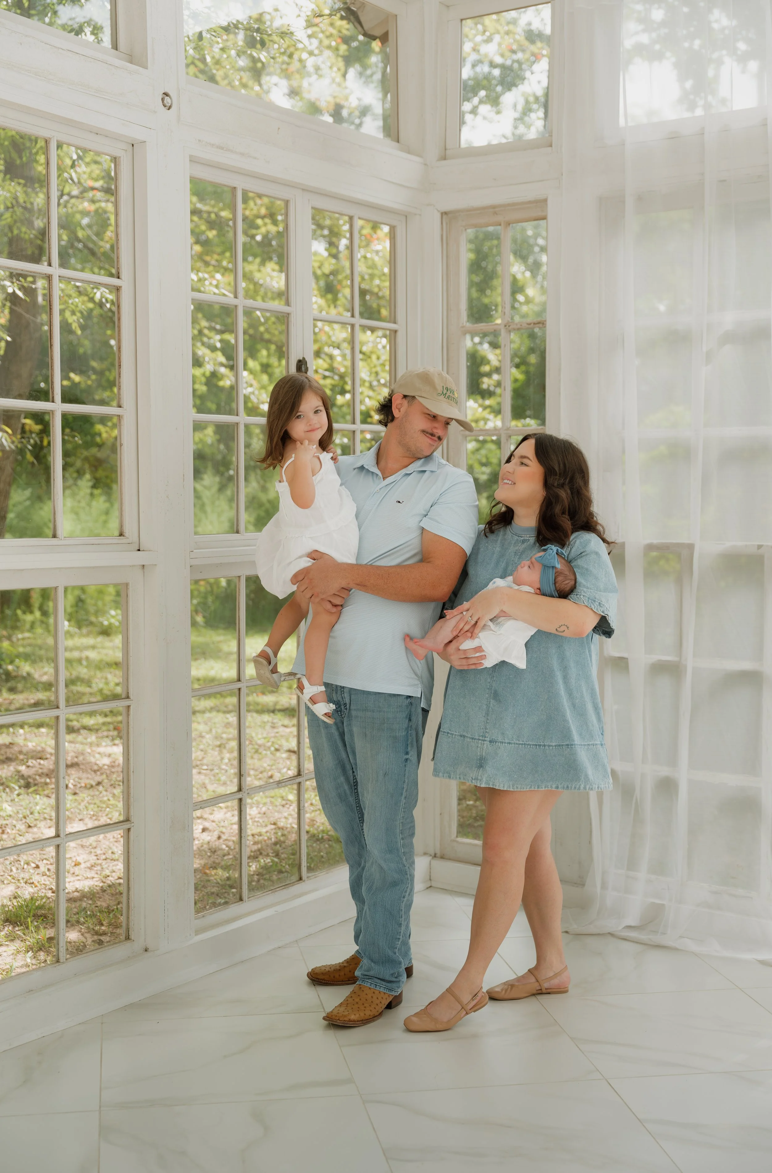 A family of four standing inside a sunlit room with large windows, holding their baby and toddler children, smiling and looking at each other.