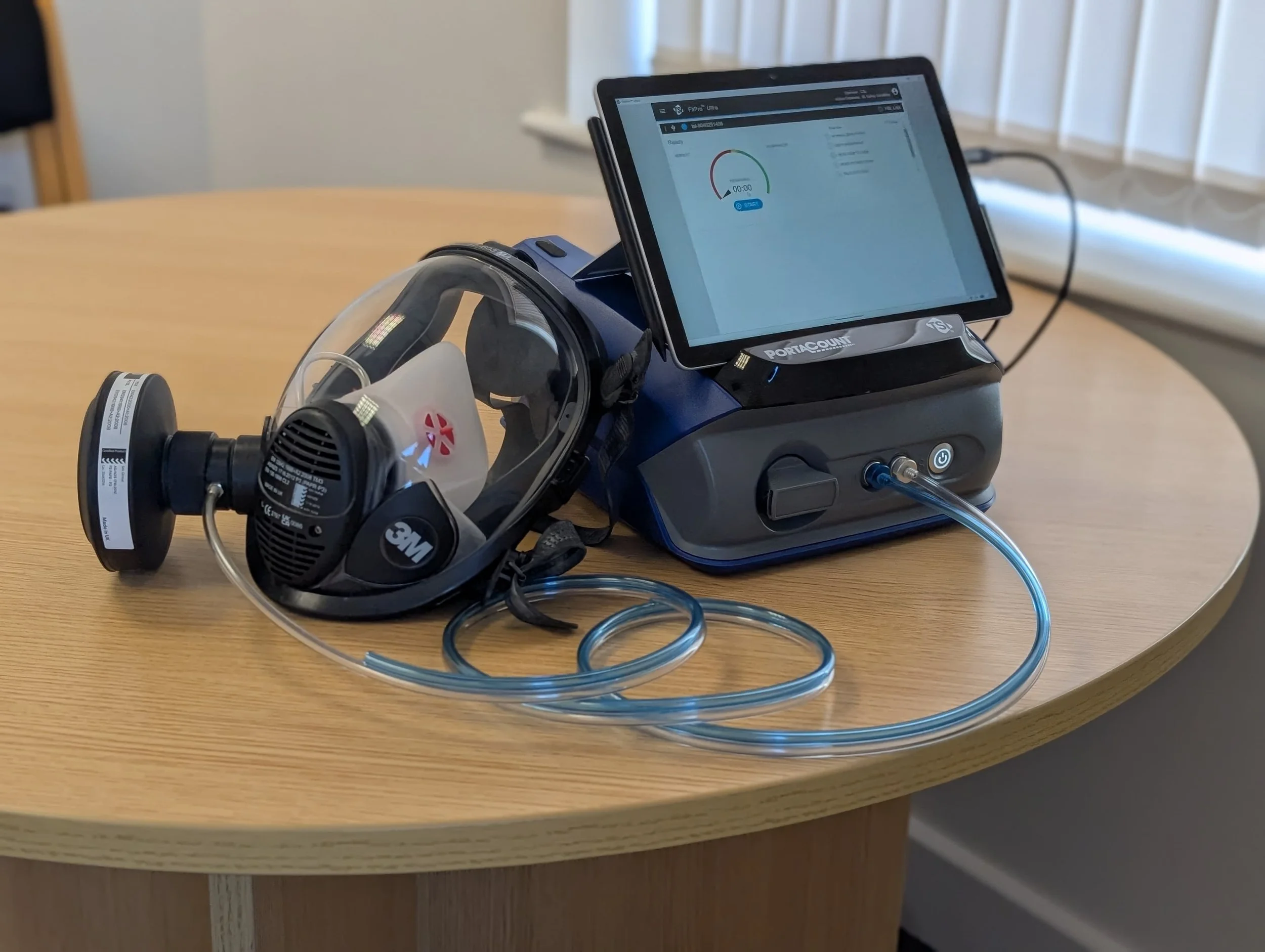 A respirator mask resting on a portacount device connected to a digital screen displaying a graph and measurements on a wooden table.