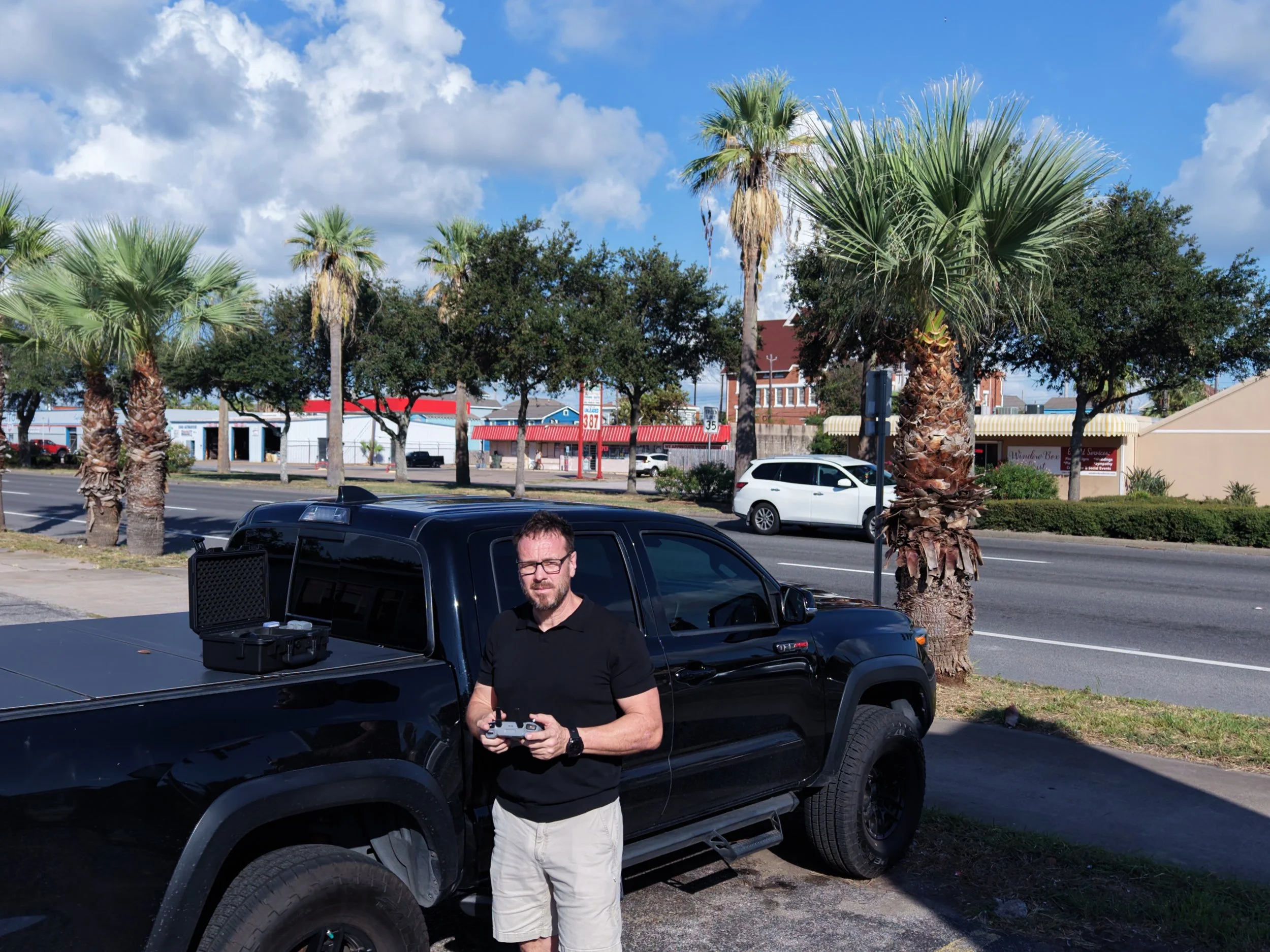 A man with glasses, a black shirt, and beige shorts holding a drone controller stands beside a black pickup truck in a parking lot. There are tall palm trees, other trees, buildings, and cars in the background under a partly cloudy sky.