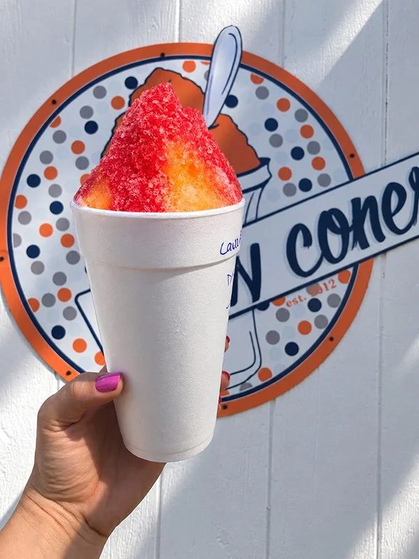 A hand holding a cup of shaved ice with red and yellow syrup, in front of a cone logo sign.