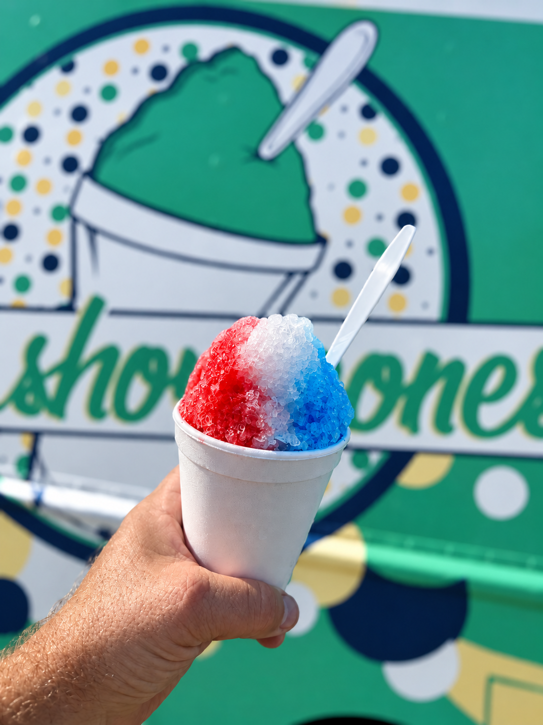 Hand holding a snow cone with red, white, and blue syrup in front of a colorful sign for a snow cone stand.