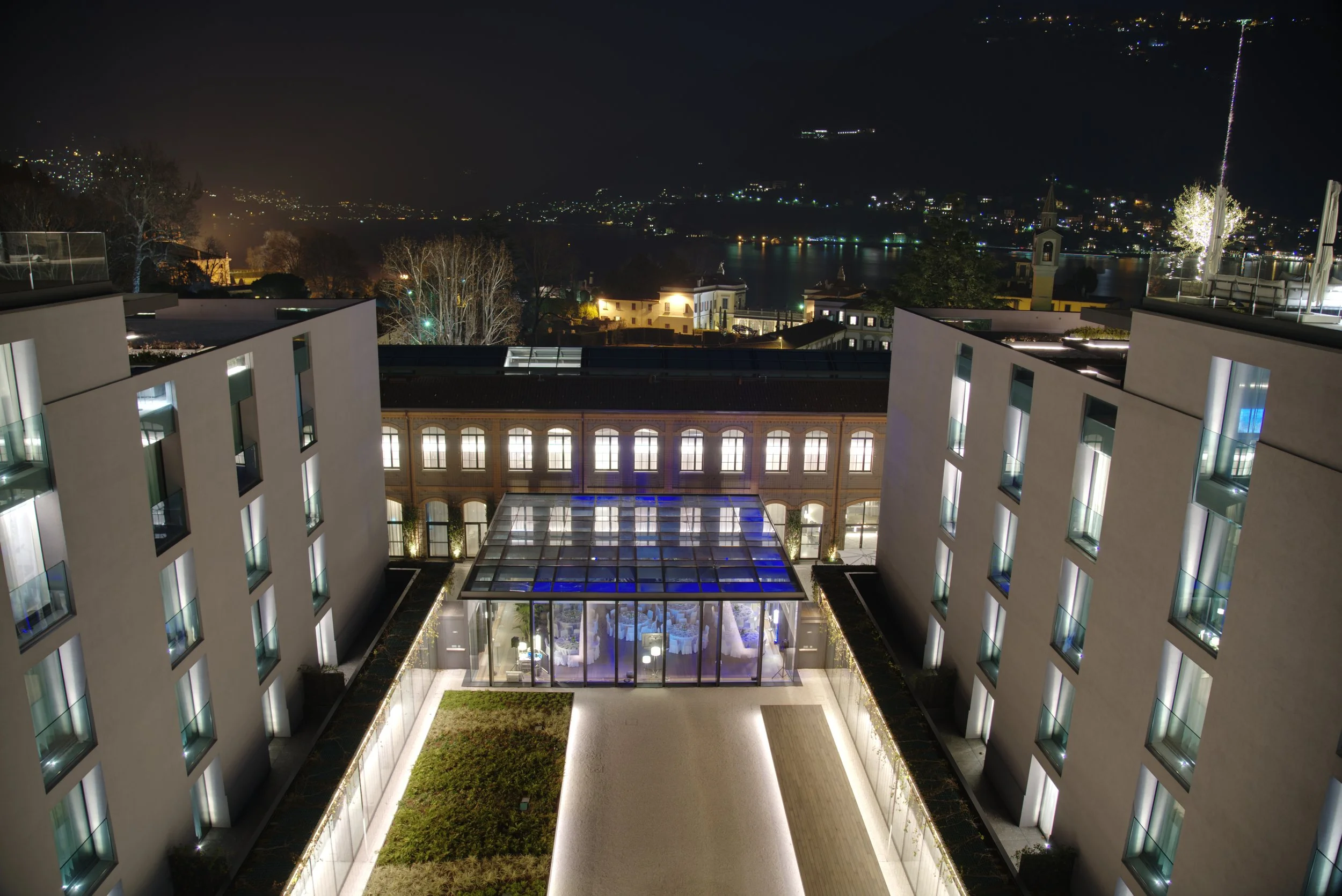 Night view of a modern hotel courtyard with illuminated windows, glass-covered walkway, and surrounding trees, overlooking a lake and a hillside town with lights.