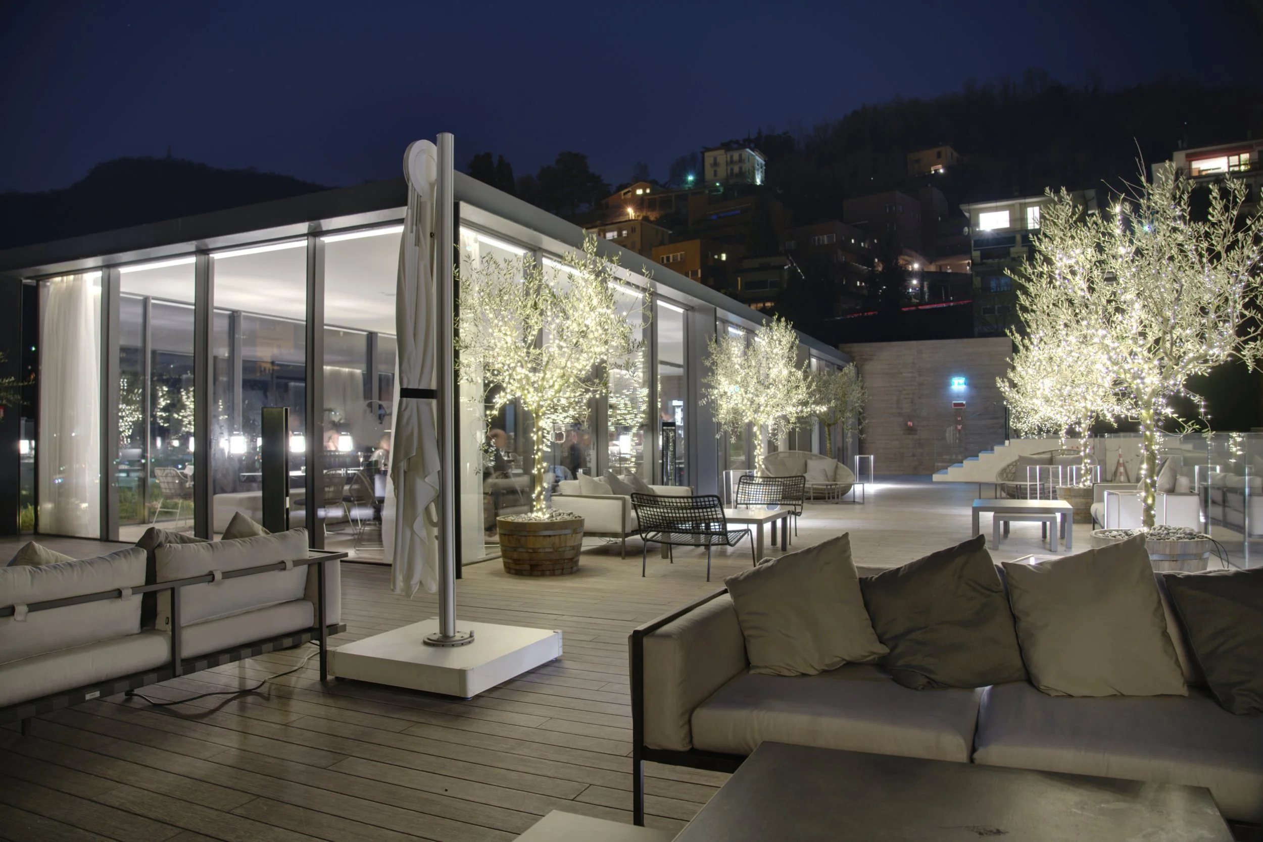Night view of an outdoor patio with modern furniture, string-lit trees, and large glass windows, with houses on a hillside in the background.